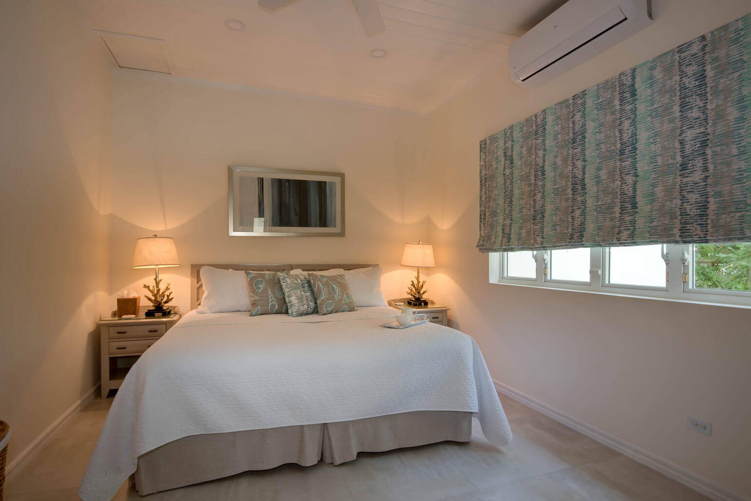 This cozy bedroom in a luxury four-bedroom holiday villa features a large bed with white linens and decorative pillows. Two bedside tables with matching lamps flank the bed, while patterned blinds dress the window. An air conditioning unit sits above, and a mirror elegantly hangs over the bed.