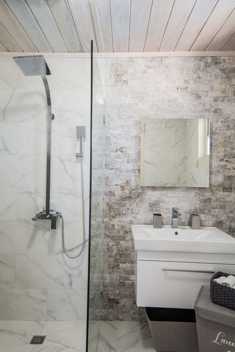 Experience the elegance of a modern bathroom found in a luxury holiday villa in Barbados, featuring a corner glass shower with a square rainfall showerhead and handheld sprayer. The marble and stone-tiled walls complement the white sink beneath a square mirror, while wood-paneled ceilings add warmth.