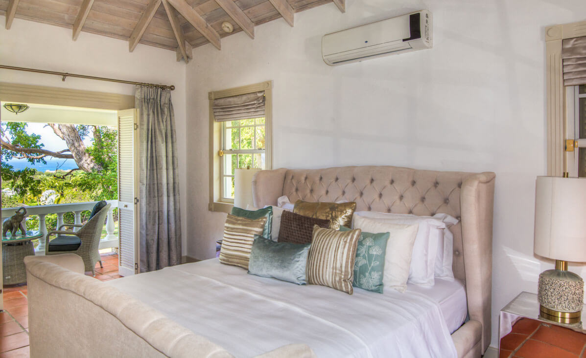 A cozy bedroom in a luxury holiday villa in Barbados features a plush, upholstered bed with decorative pillows. A window and open doorway leading to a balcony showcase views of greenery and ocean. The room has a pitched wooden ceiling and warm lighting, creating a tranquil atmosphere.