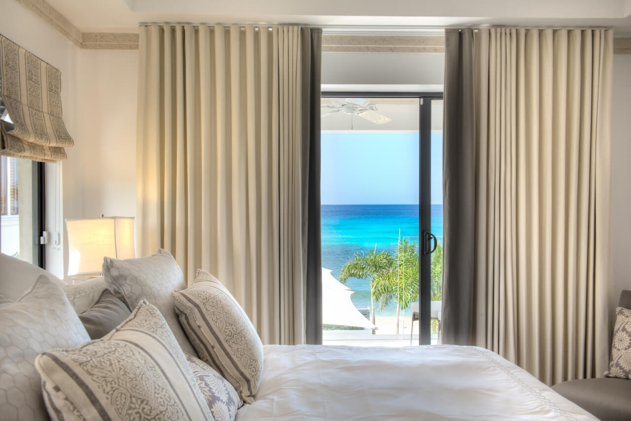 A luxurious bedroom in a contemporary beachfront villa offers a stunning ocean view through large sliding glass doors. The room features elegant beige curtains, a plush bed with decorative pillows, and a soft lamp on the nightstand, with palm trees and blue water visible outside.
