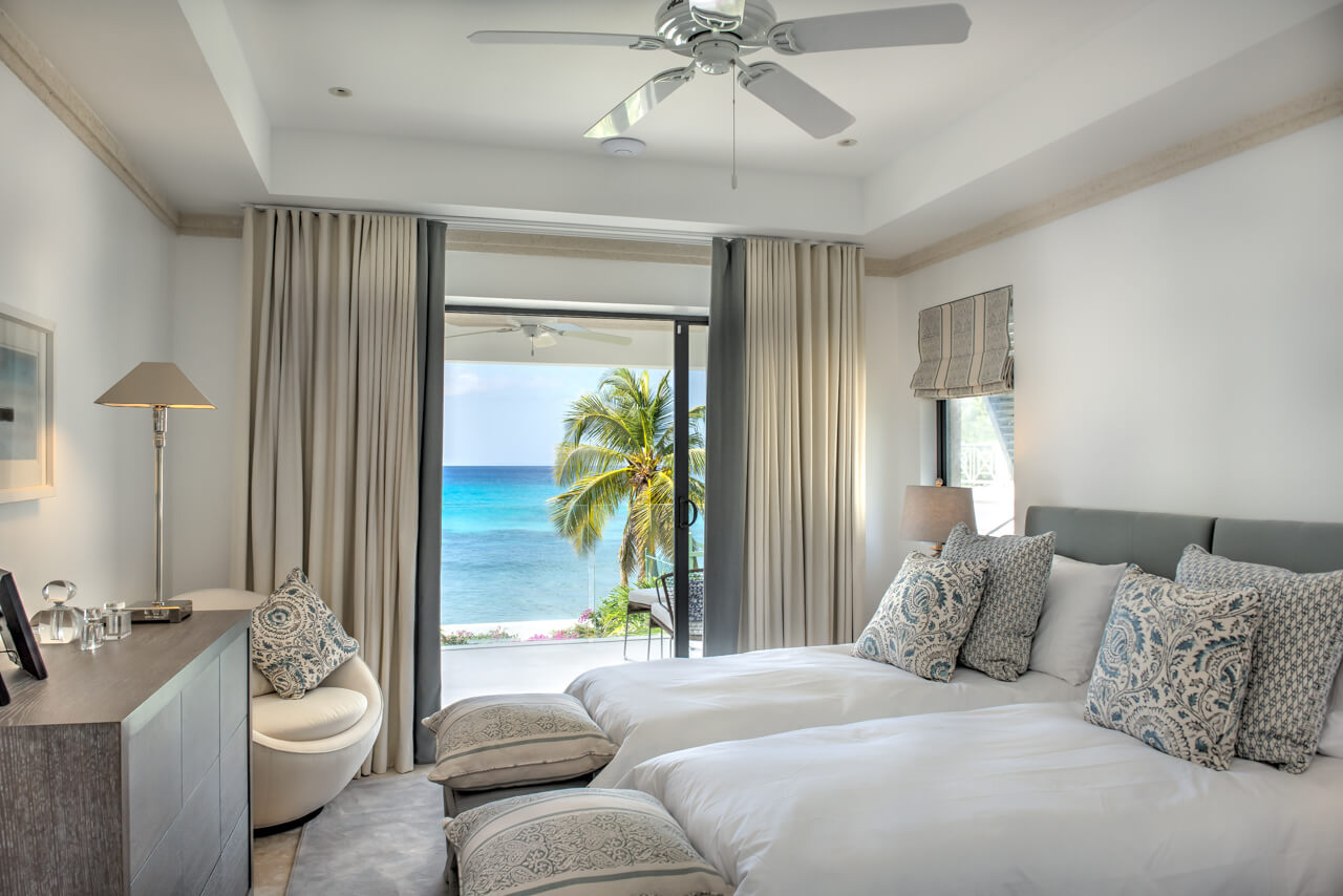 A serene bedroom in a contemporary luxury beachfront villa features twin beds adorned with patterned pillows, facing a sliding glass door. This door opens to a balcony overlooking a palm tree and the ocean under clear blue skies, with light curtains and a ceiling fan enhancing the breezy feel.