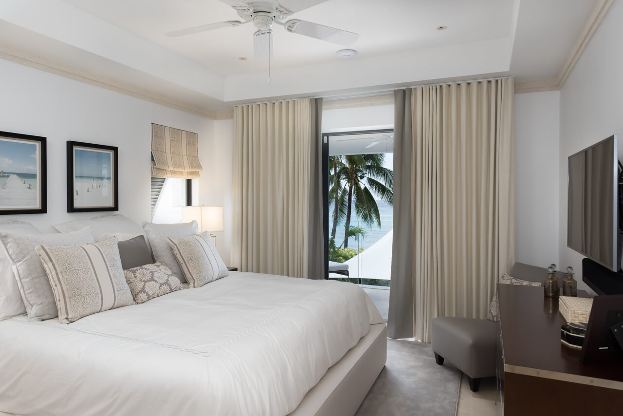 A serene bedroom in a contemporary luxury beachfront villa features a large bed with patterned pillows and framed artwork. Curtains open to reveal palm trees and the ocean, while soft lighting and a ceiling fan enhance the tranquil ambiance. A TV rests on a wooden dresser.