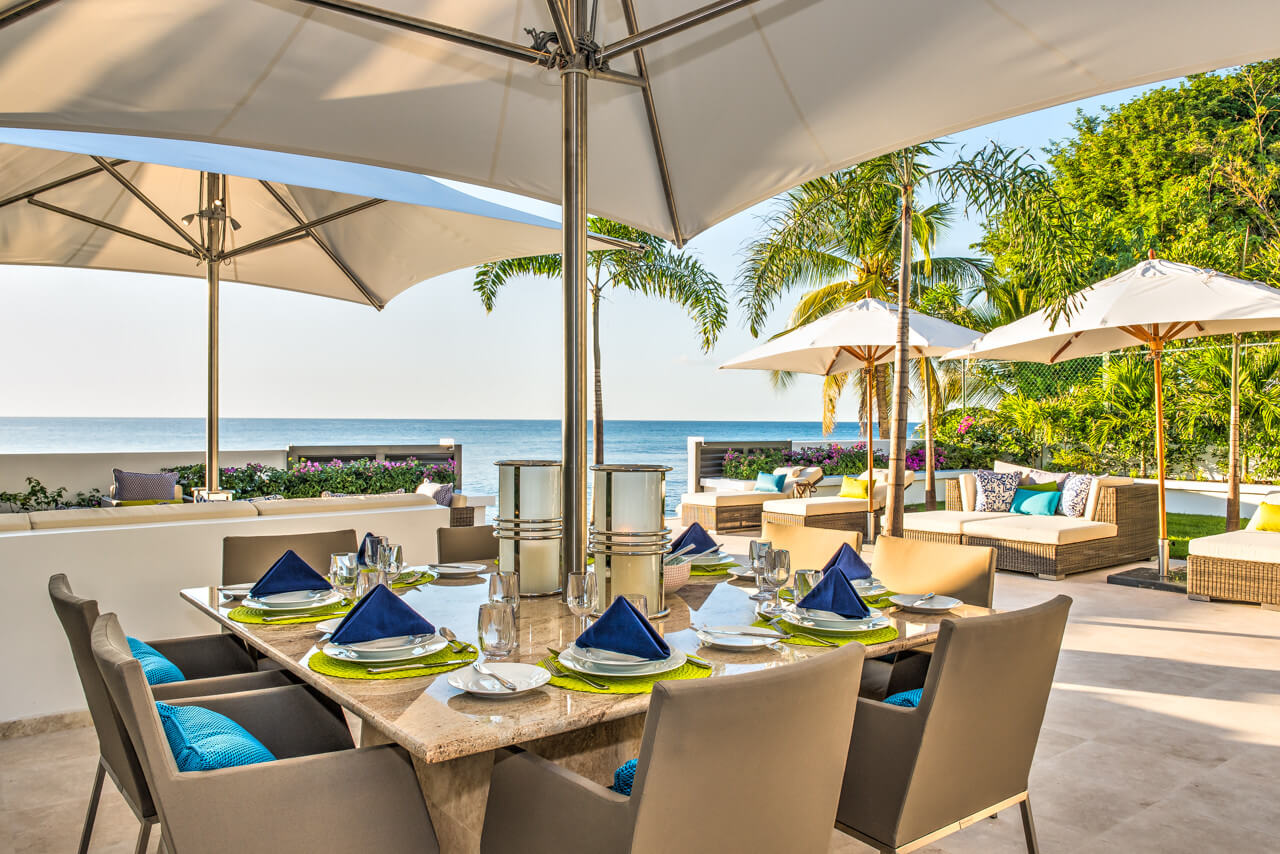 The contemporary luxury beachfront villa boasts an outdoor dining area by the ocean, featuring a set table under large umbrellas. Surrounded by palm trees and cushioned seating, the scene is enhanced with blue napkins, exuding a sunny and tranquil atmosphere.