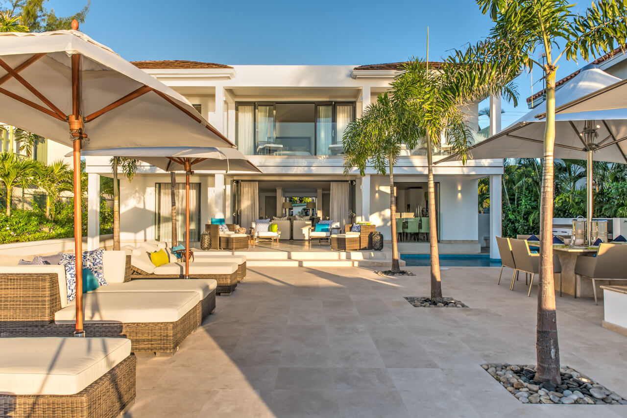 Contemporary luxury beachfront villa boasting a white exterior and large glass windows, with a spacious patio featuring cushioned seating, umbrellas, palm trees, and an outdoor dining area under the clear blue sky.
