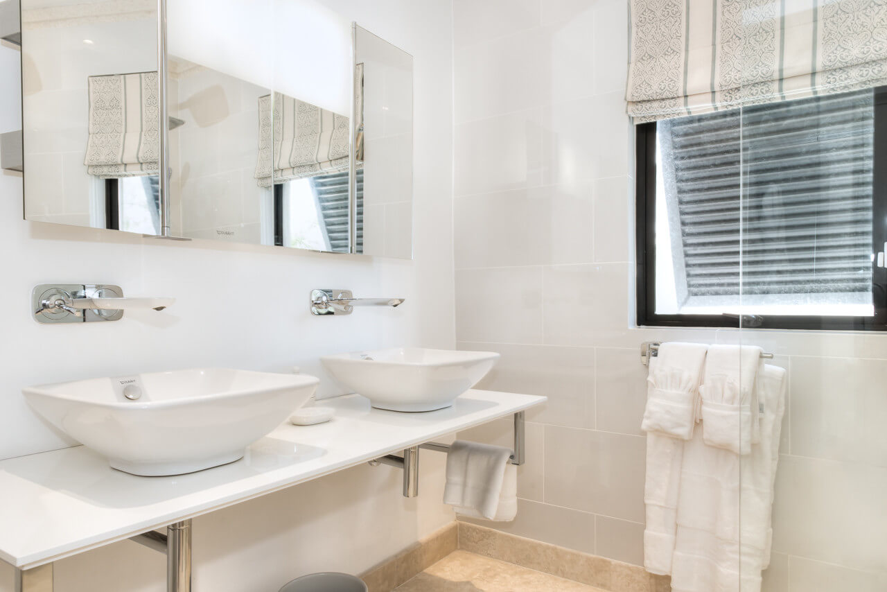 A contemporary bathroom in a luxury beachfront villa features two rectangular white sinks with wall-mounted faucets on a sleek countertop. Mirrors hover above the sinks, while a window with striped blinds invites natural light. Towels are elegantly draped on a rack below the counter.
