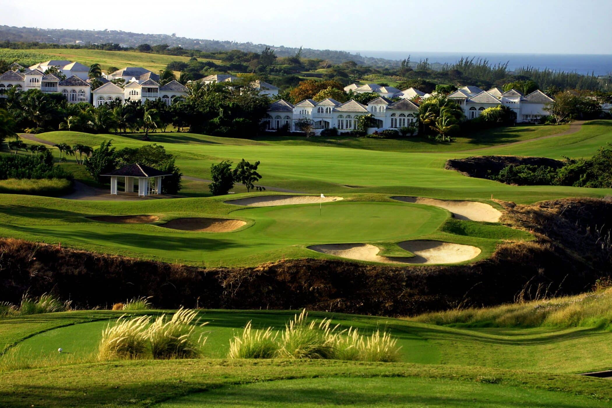 A scenic view of a lush golf course with neatly trimmed greens and sand bunkers, bordered by elegant white villas and surrounded by greenery under a clear sky.