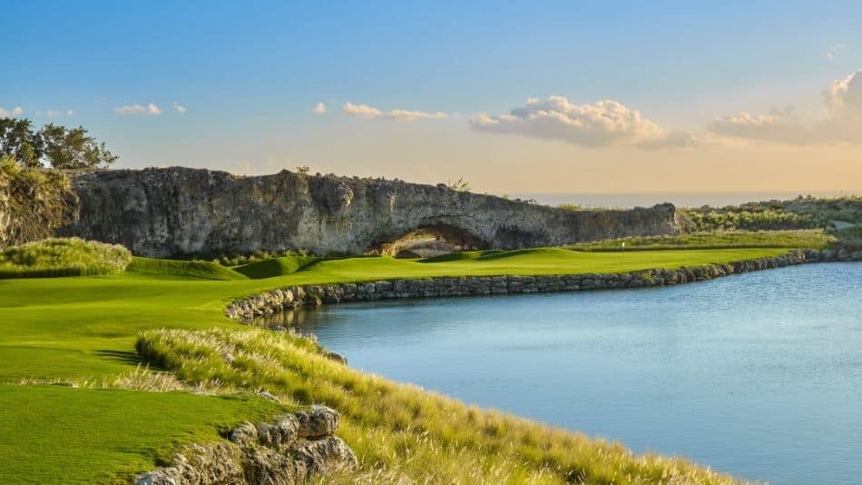 A scenic golf course with lush green fairways beside a calm blue water hazard, bordered by rocky terrain and tall grass, under a clear sky with soft clouds at sunset.