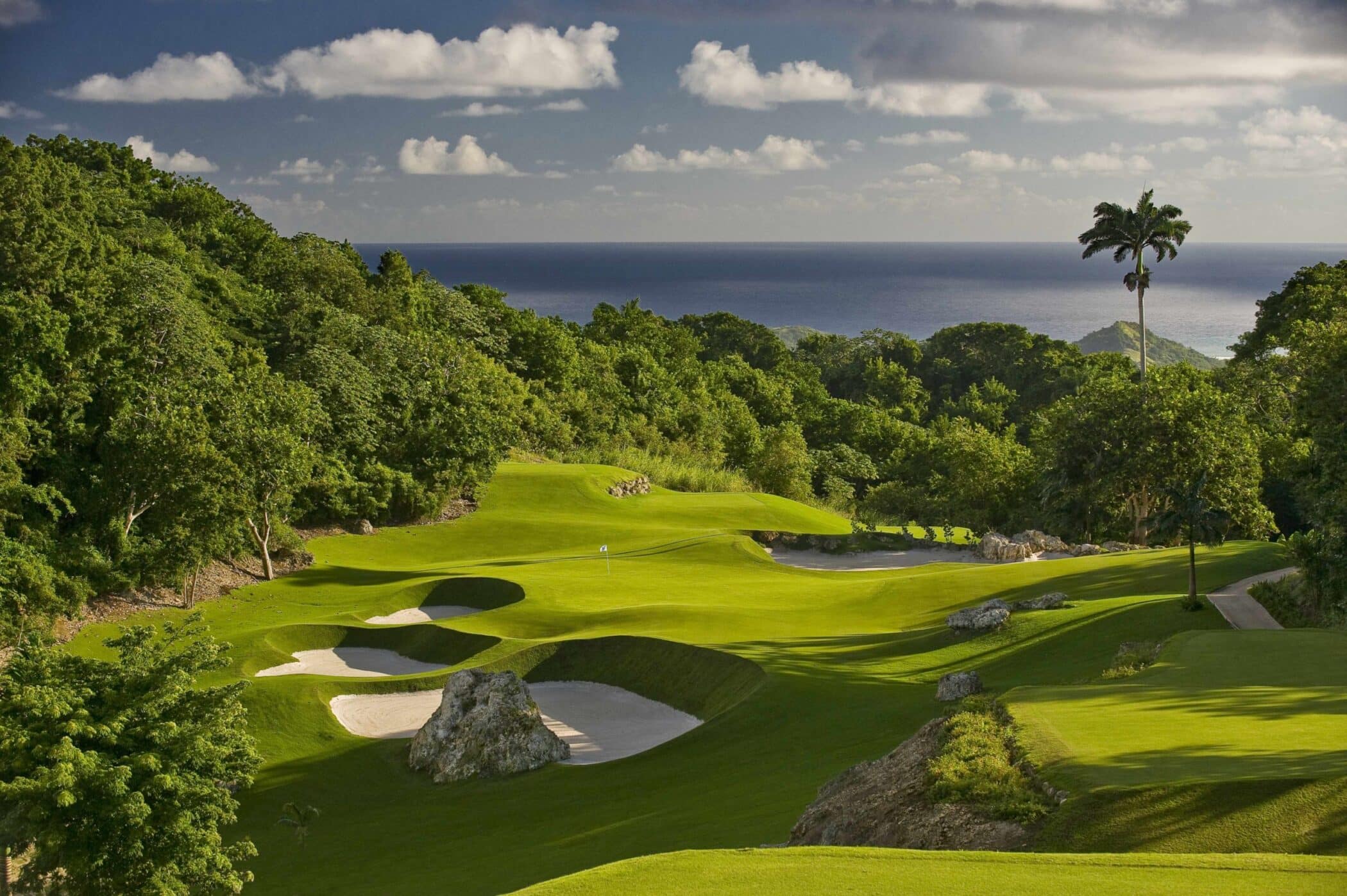 A scenic golf course with sand bunkers, lush green fairways, and tropical trees overlooking the ocean under a partly cloudy sky.