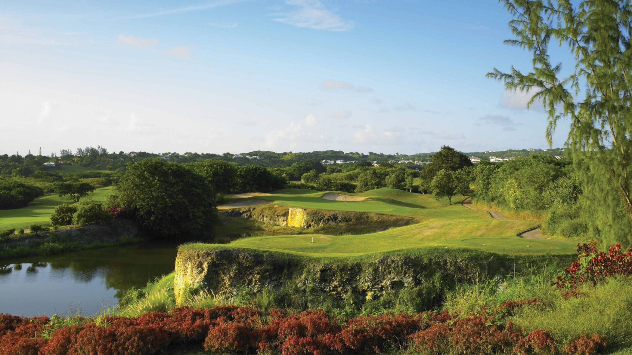 A lush, green golf course with sand bunkers, a pond, and rocky edges, surrounded by trees and bushes under a clear blue sky with distant houses in the background.