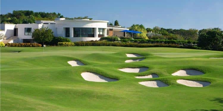 A modern white building overlooks a lush green golf course with numerous sand bunkers and manicured grass under a clear blue sky. Trees and shrubs surround the area.