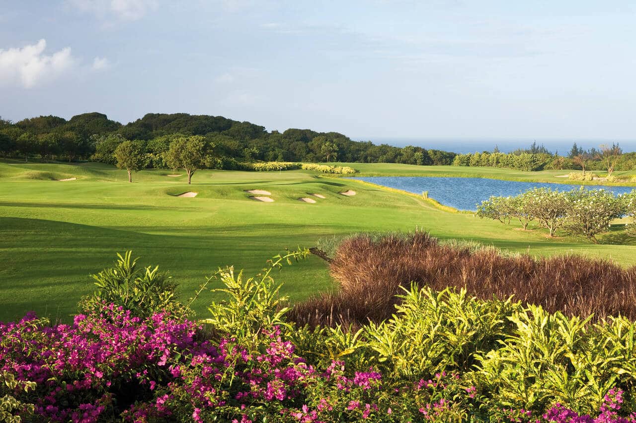 A scenic golf course with lush green fairways, sand bunkers, and a pond, surrounded by vibrant flowering bushes and trees under a clear blue sky.