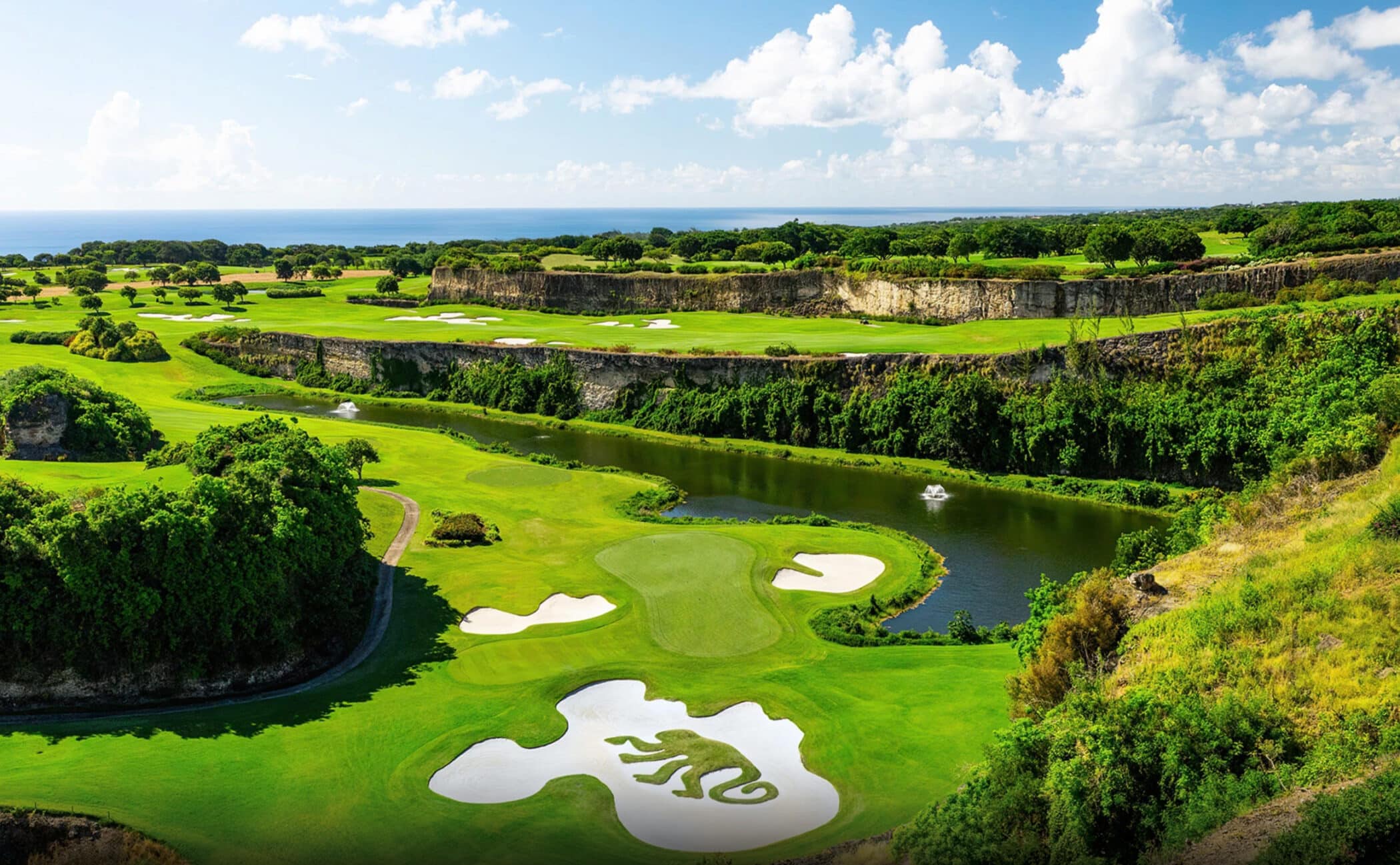 A scenic golf course featuring lush green fairways, white sand bunkers, water hazards, and elevated cliffs under a bright blue sky with scattered clouds. The ocean is visible in the distant background.