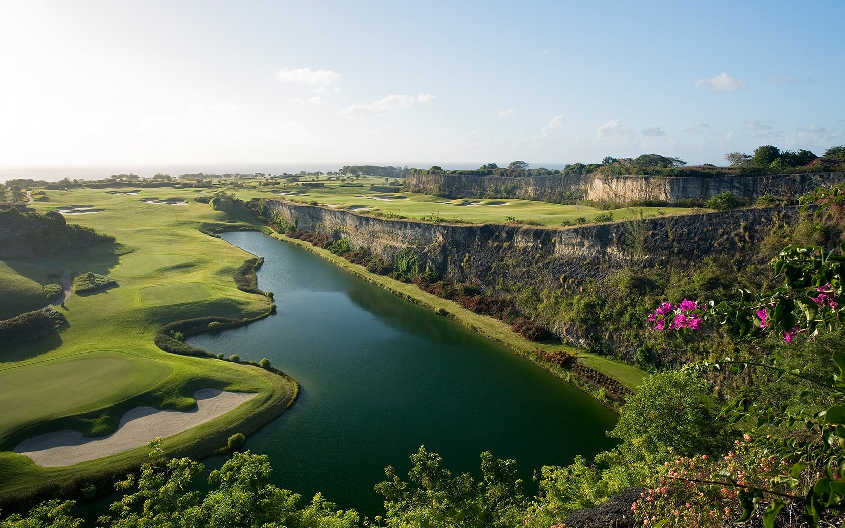 A scenic view of a golf course with green fairways, sand bunkers, a river running through the landscape, high cliffs, and flowering bushes under a blue sky.