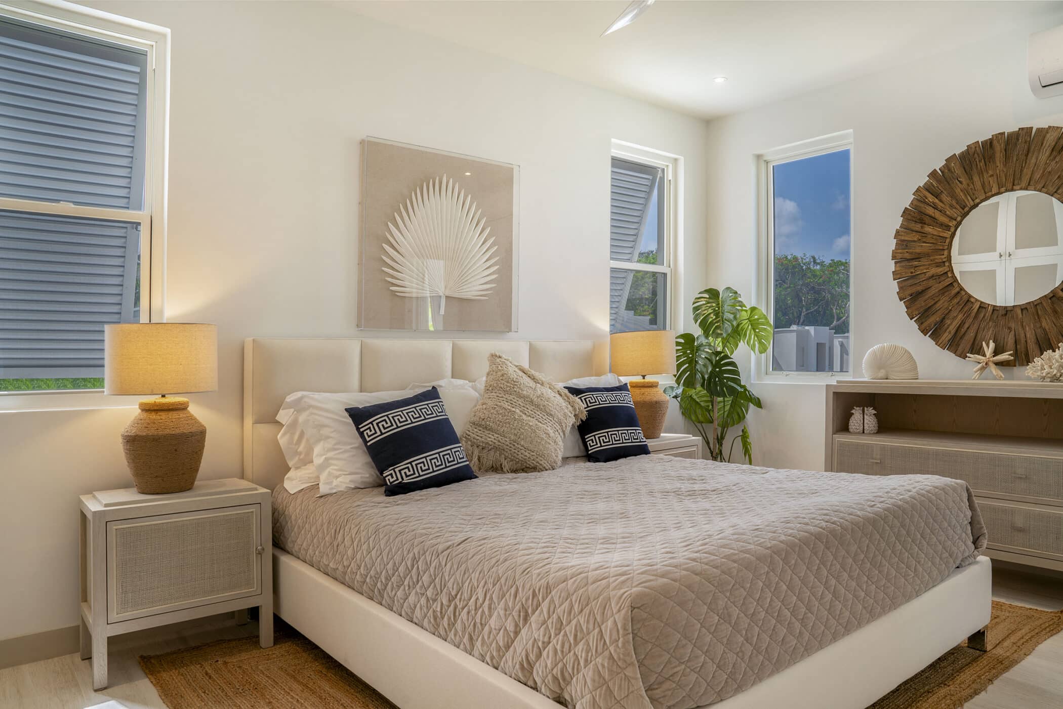 A cozy bedroom at The White House on Mango Drive features a beige quilted bed, two bedside tables with lamps, tropical decor, a lush plant, and a dresser with a round mirror. Sunlight streams through three windows.
