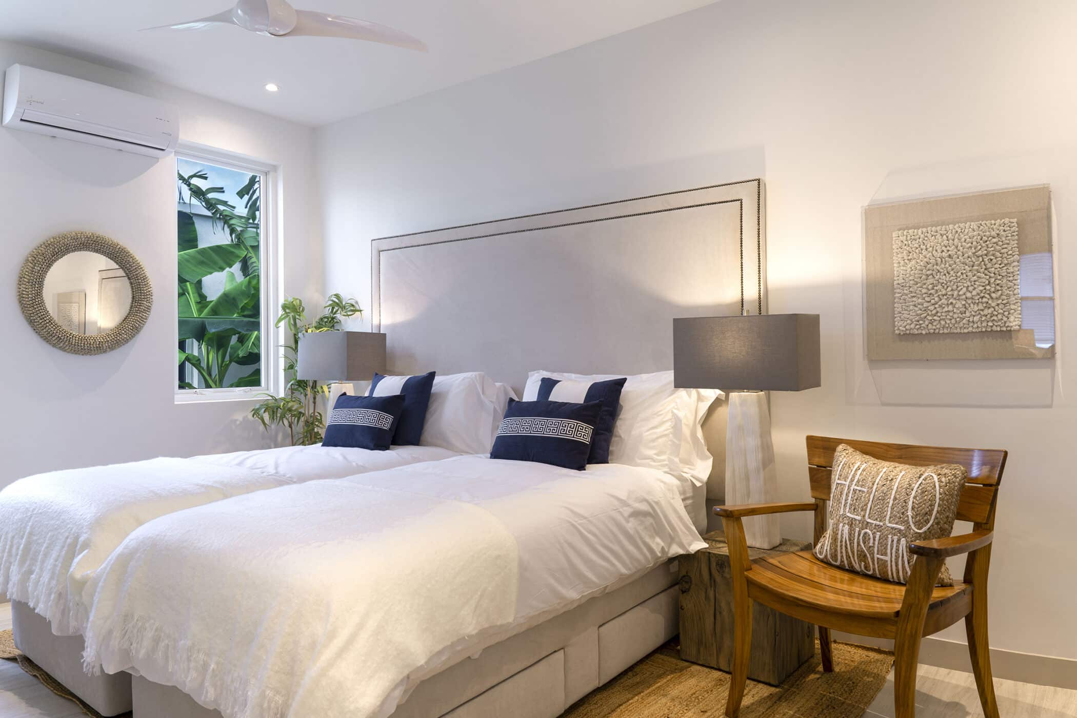 Bright bedroom with two twin beds, white bedding, and navy accent pillows at The White House on Mango Drive. The space features two gray lamps, a wooden chair with a “Hello Sunshine” pillow, wall art, a round mirror, and a window revealing lush green plants outside.