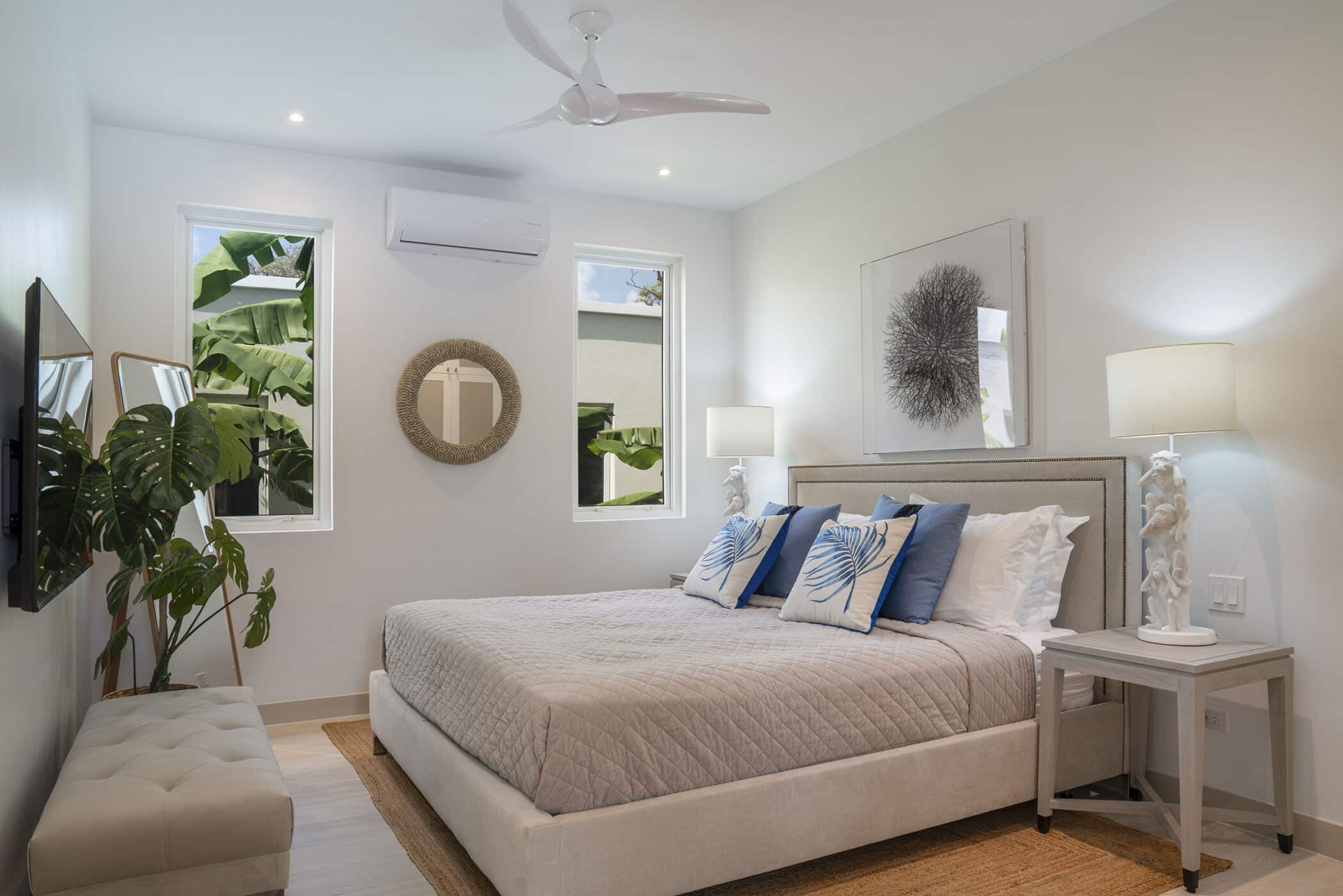 Modern bedroom in The White House on Mango Drive with a beige bed, blue and white pillows, two bedside tables with lamps, wall art, a TV, a large plant, and two windows letting in natural light.