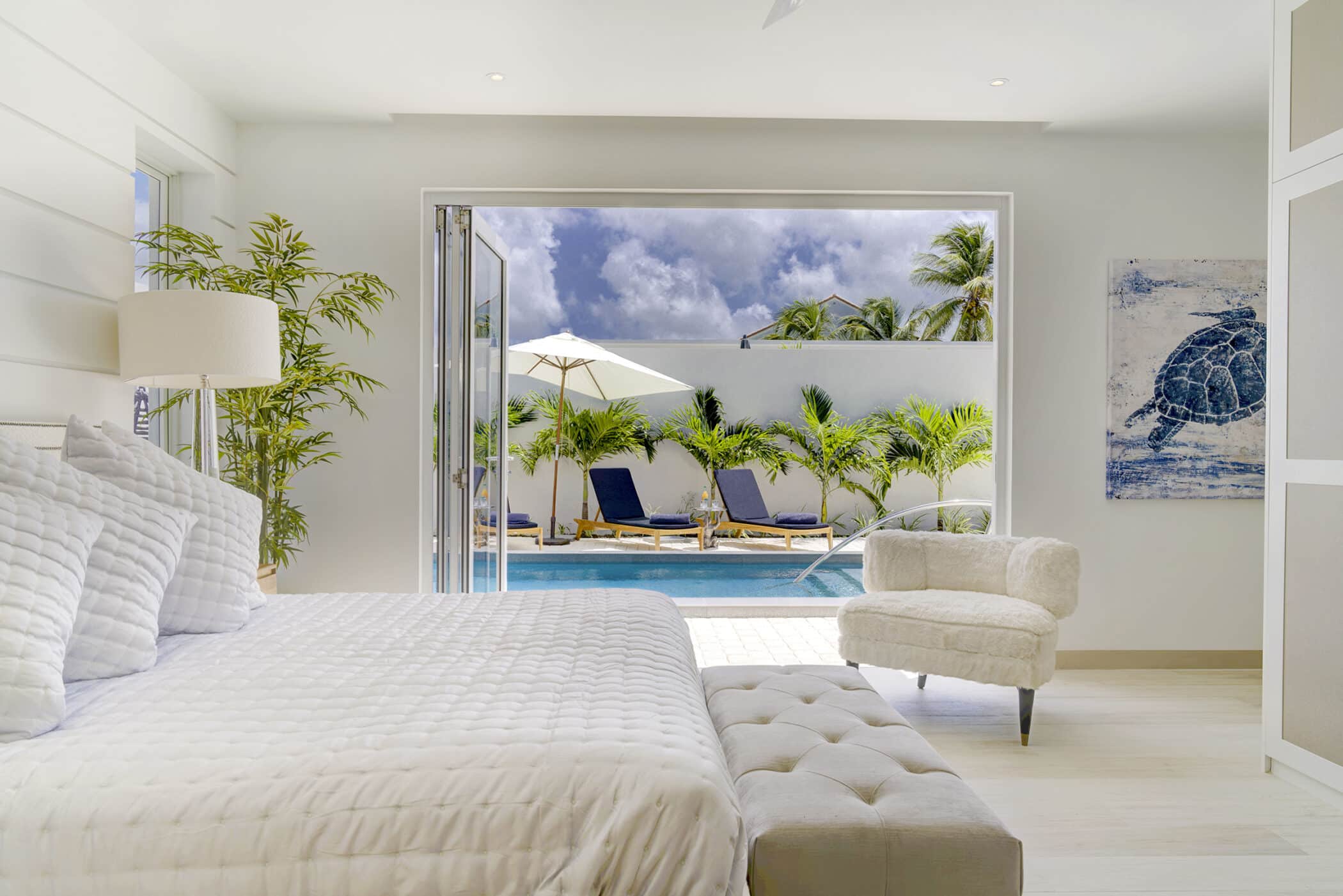 A bright bedroom at The White House on Mango Drive features a white bed and chair, opening to a pool area with lounge chairs, umbrellas, palm trees, and a blue sky dotted with clouds.