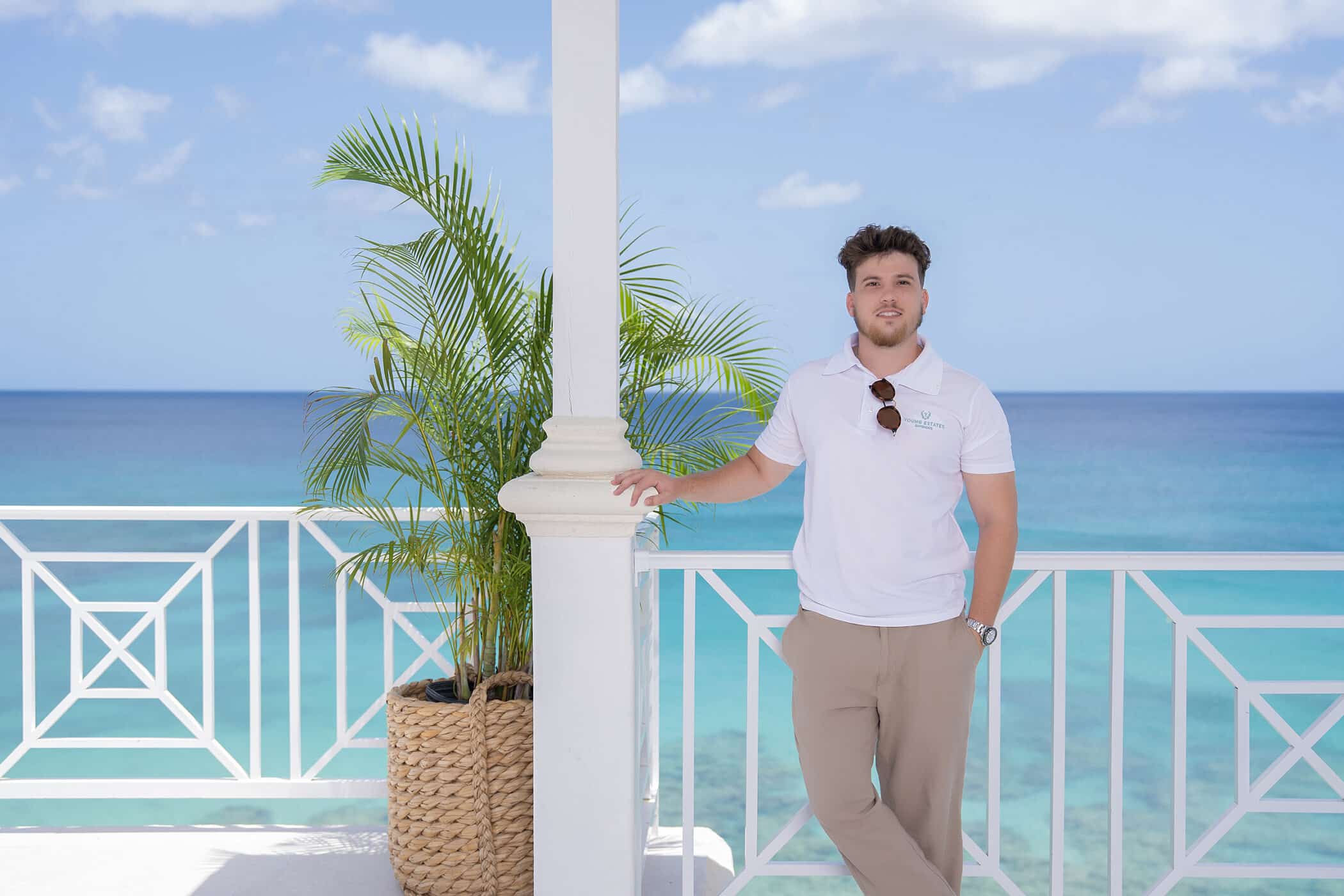 A man in a white polo shirt and beige pants stands by a white railing on a balcony at Beachi at Coral Cove, overlooking a turquoise ocean, with a potted palm nearby and a clear blue sky in the background.