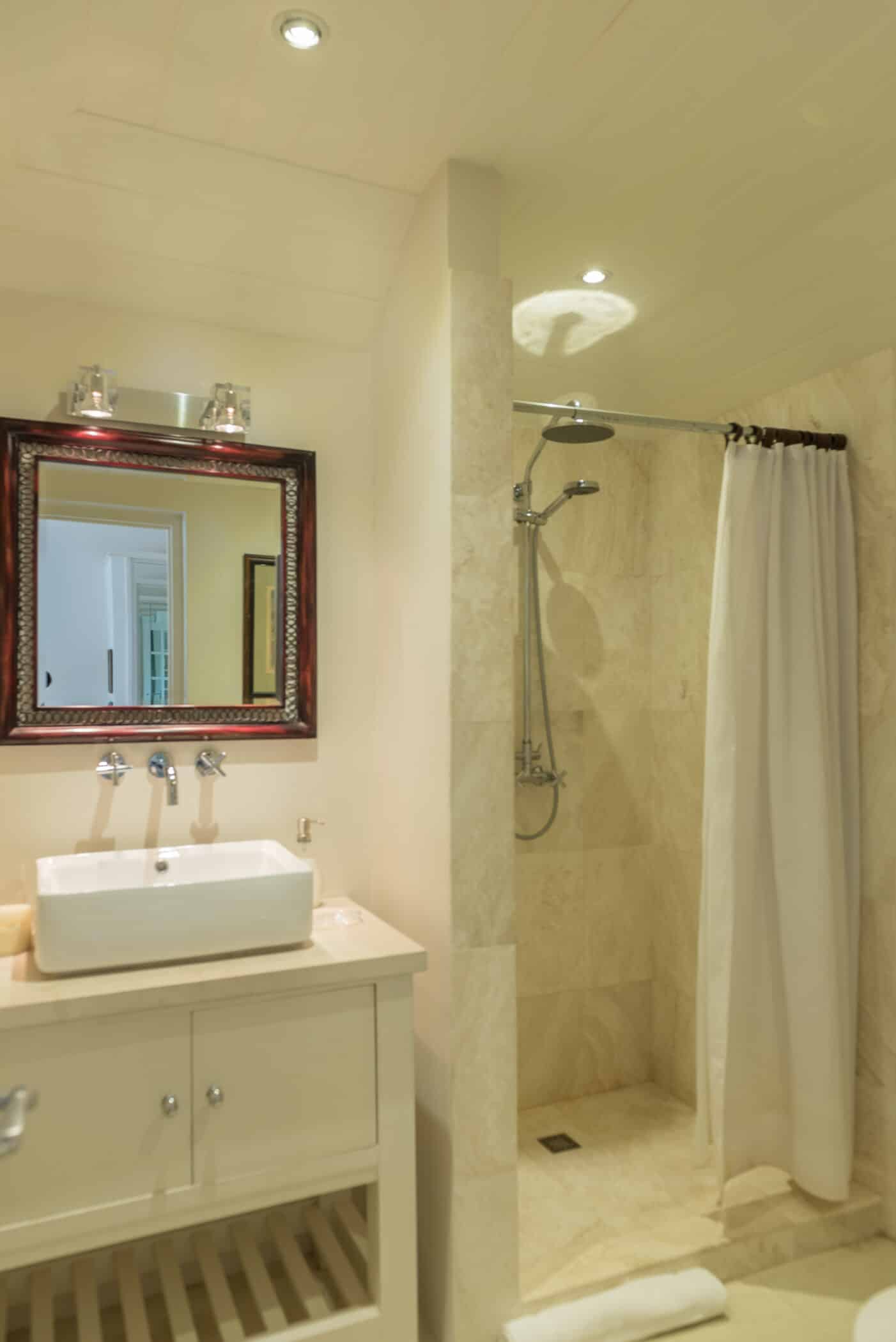 This bathroom at Casablanca at Sandy Lane features a white vanity, rectangular sink, wall mirror, and a walk-in shower with beige tiles and a white curtain. The softly lit space is enhanced by recessed and wall lights.