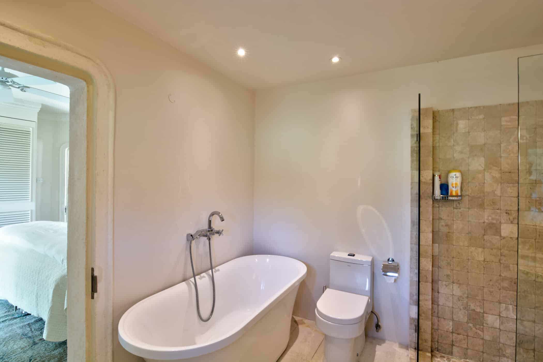 Modern bathroom with a freestanding bathtub, a toilet, and a glass-enclosed shower with beige tiles at Casablanca at Sandy Lane. A bedroom with a white bed is visible through an open door. The light-colored walls and recessed ceiling lights add to the contemporary elegance of this luxurious space.