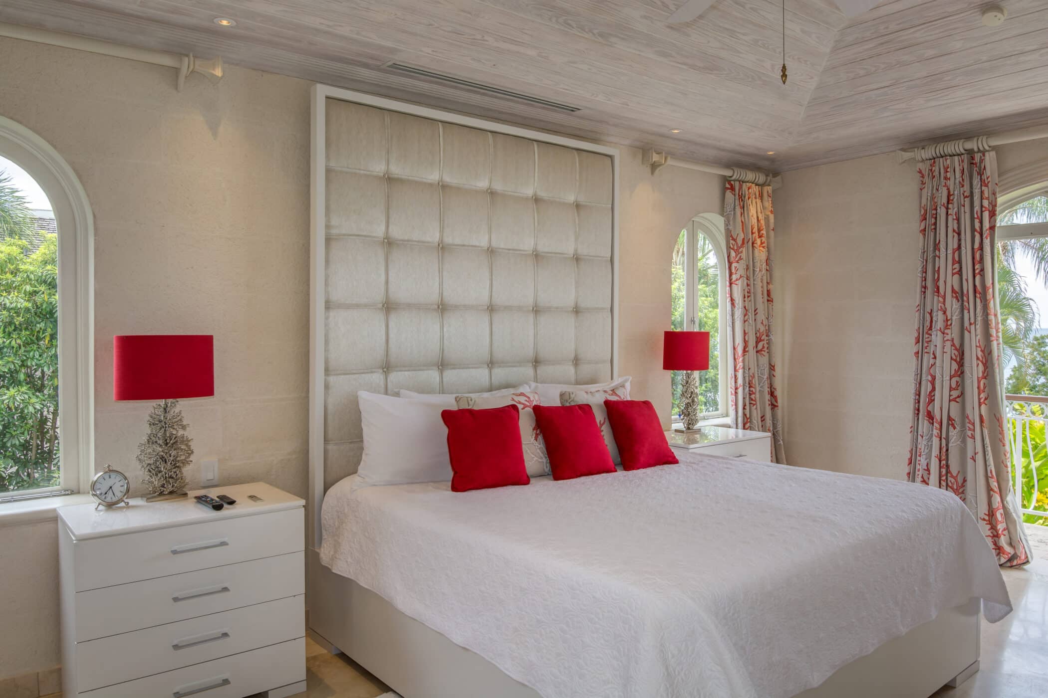 A bright bedroom at Firefly at Old Trees features a large bed with crisp white bedding, red accent pillows, matching red lamps, two nightstands, and elegant arched windows adorned with curtains. Lush greenery is visible outside through the windows.