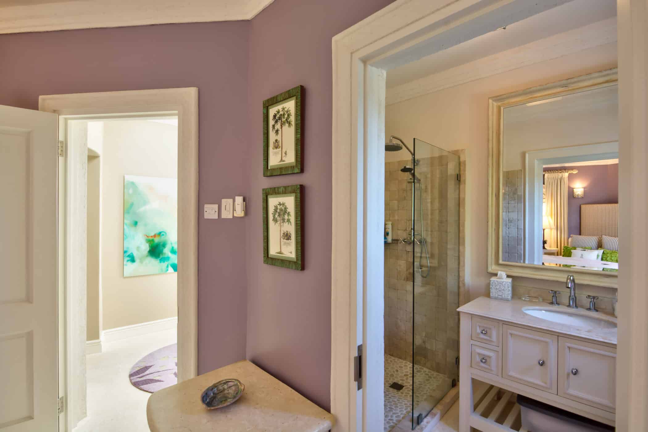 A bathroom with lavender walls, two framed botanical prints, and a beige counter. Through an open door at Casablanca at Sandy Lane, see a vanity with a mirror, a glass shower, and a glimpse of a bed and abstract art in another room.