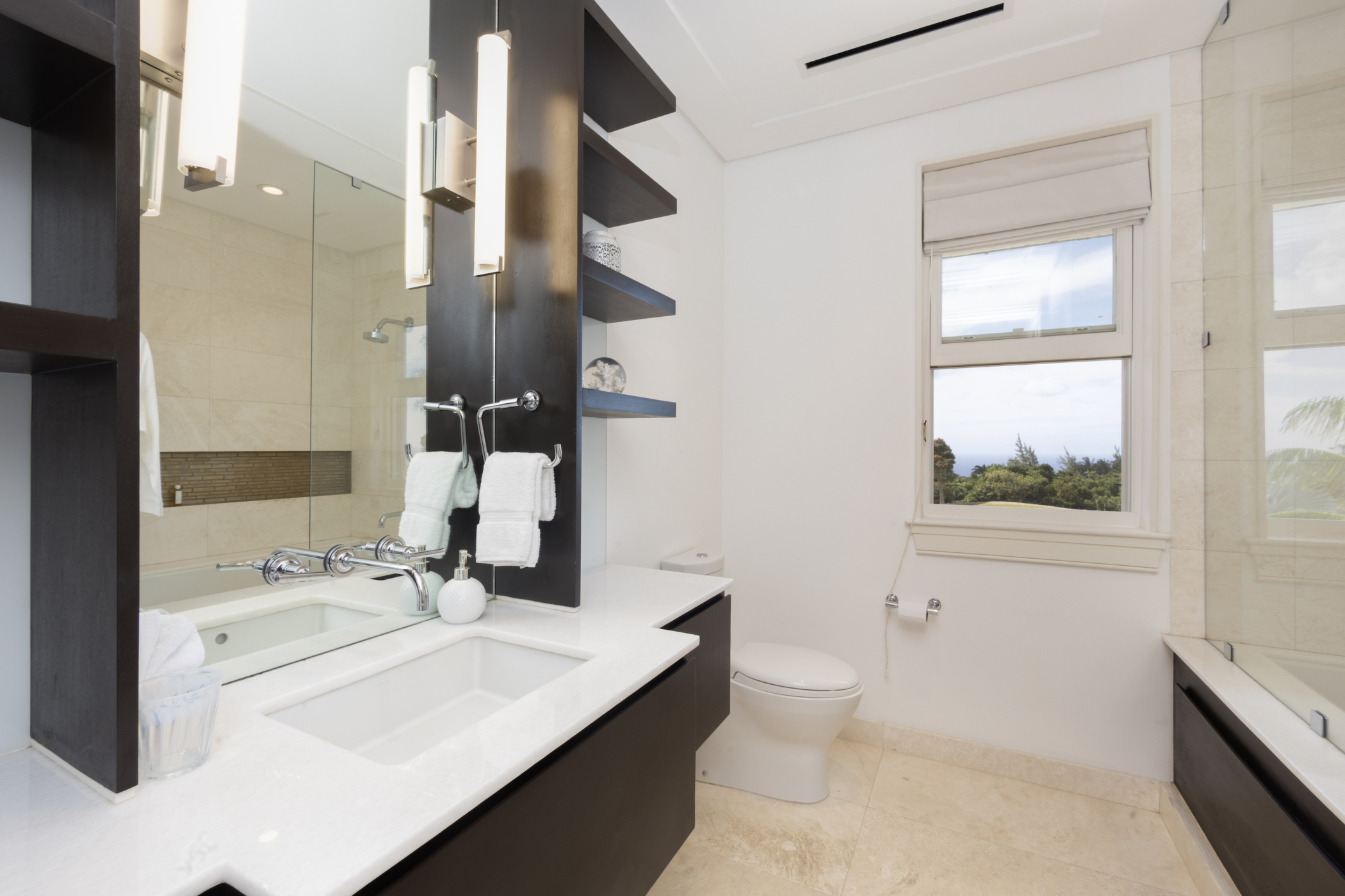 Modern Cocomaya bathroom with a white countertop, sink, mirror, shelves with towels, and a toilet next to a window. The window offers a view of trees and sky outside. The space is bright and clean.