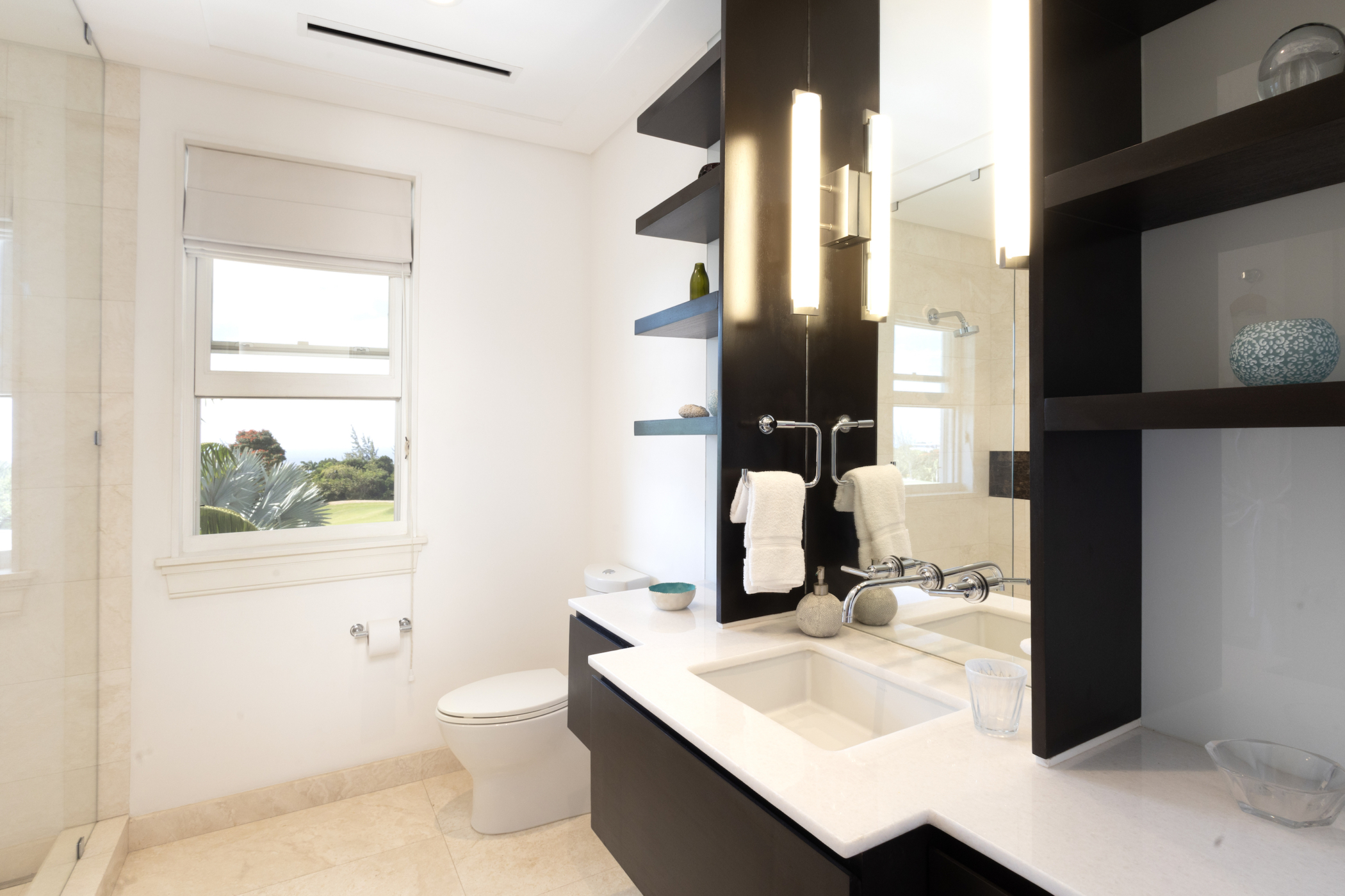 Modern Cocomaya bathroom with a white countertop, sink, toilet, and large mirror. Built-in shelves hold decor items, and a window lets in natural light. Wall-mounted lights add brightness beside the mirror.