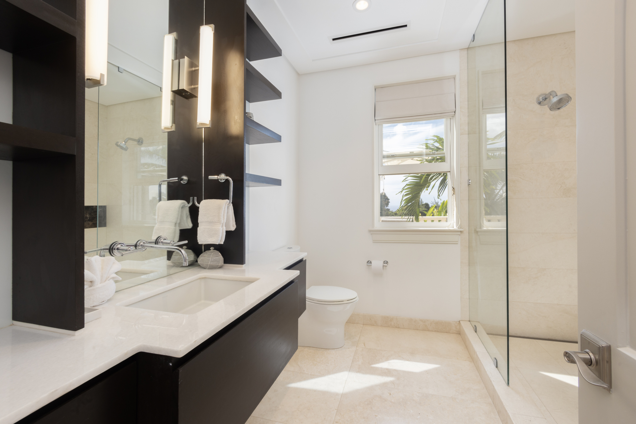 Modern Cocomaya bathroom with a glass shower, white toilet, large mirror, black shelves, white sink with chrome fixtures, and a window letting in natural light. Towels are neatly hung near the sink.