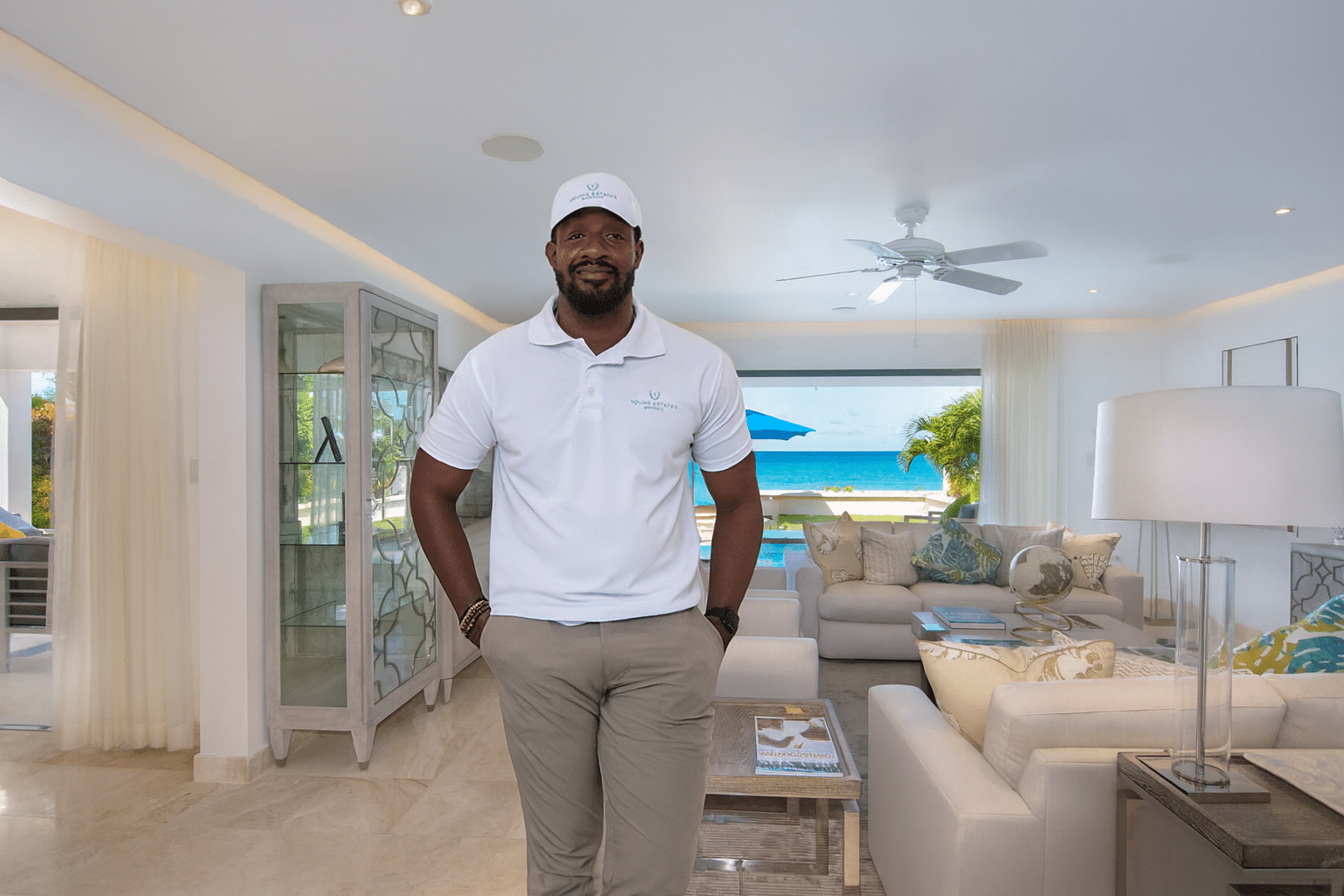 A man in a white polo shirt and cap stands smiling in a bright, modern living room with large windows revealing an ocean and palm trees outside—a true beachside nirvana, decorated in light tones and coastal accents.