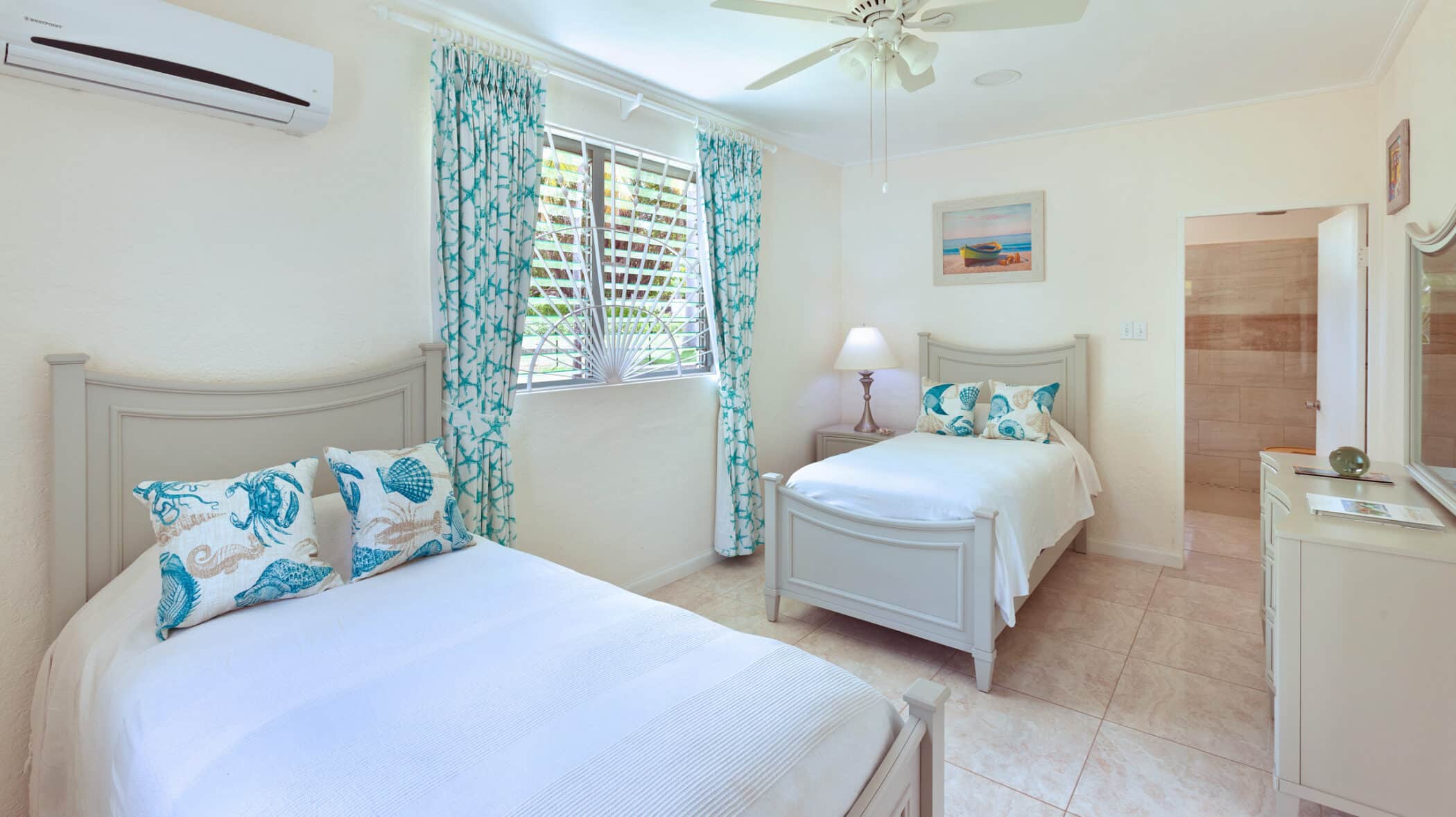 A bright bedroom with two white beds, sea-themed pillows, a bedside lamp, tropical-print curtains, a ceiling fan, a dresser with a mirror, and a window letting in natural light.