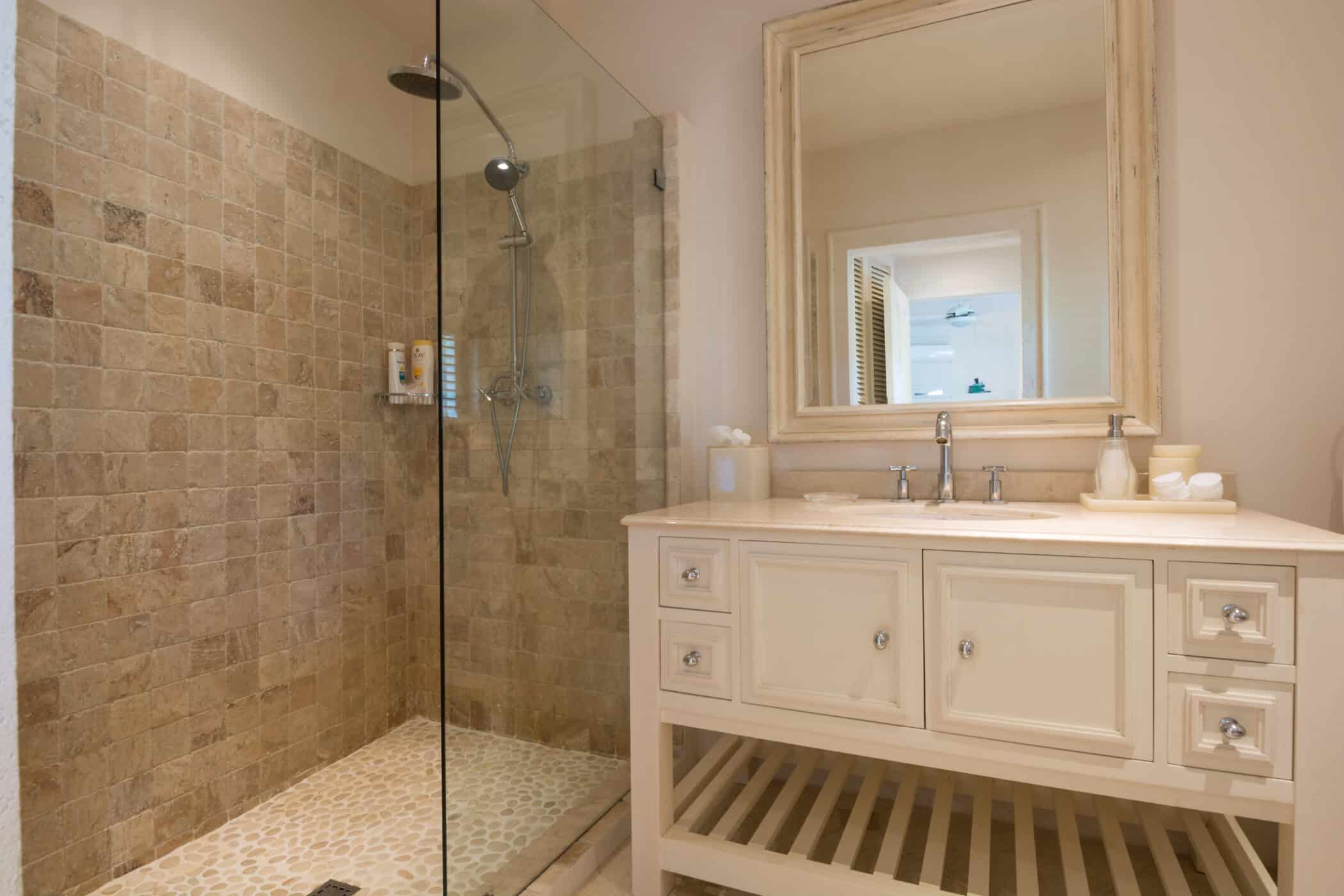 A bathroom at Casablanca at Sandy Lane features a glass-enclosed shower with beige stone tiles and a rainfall showerhead, next to a white vanity with a marble countertop, drawers, and a large framed mirror above.