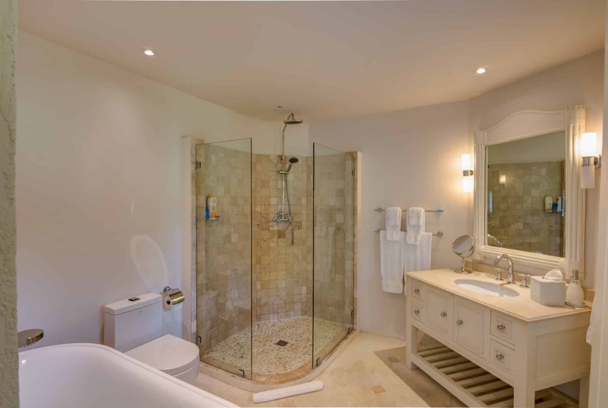 A modern bathroom at Casablanca at Sandy Lane features a corner glass shower, white toilet, freestanding bathtub, and a white vanity with sink and mirror. Soft lighting and beige stone tiles create a warm, inviting atmosphere.