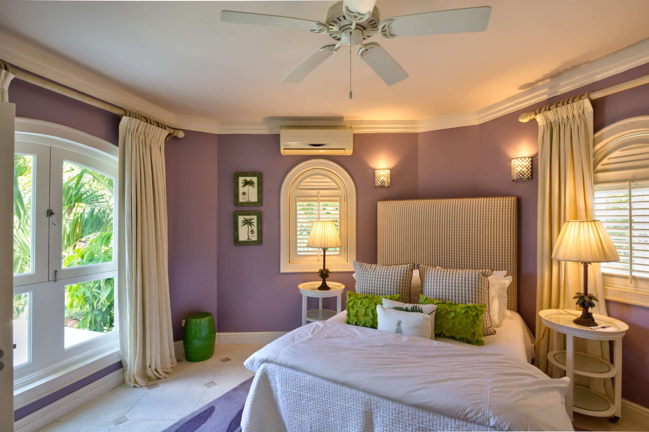 A cozy bedroom with purple walls, large windows, a bed with white and green accents, side tables with lamps, framed art, and a ceiling fan offers a welcoming retreat at Casablanca at Sandy Lane. Sunlight streams in, brightening the space and highlighting its inviting charm.