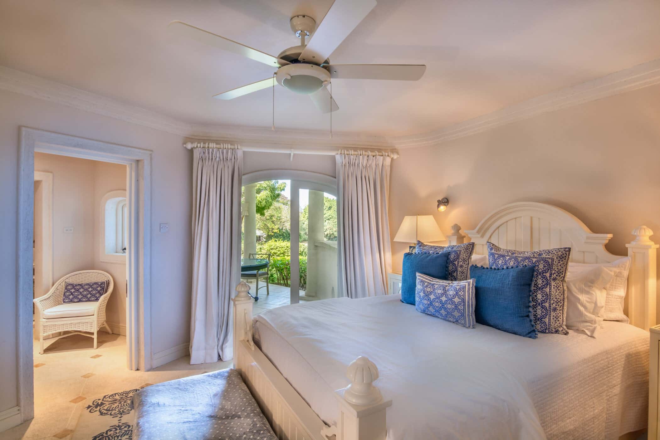 Bright bedroom with a white bed, blue accent pillows, ceiling fan, and large window doors leading to a green garden—an inviting escape at Casablanca at Sandy Lane. A chair sits in an adjacent sunlit room, and a lamp rests on the bedside table.