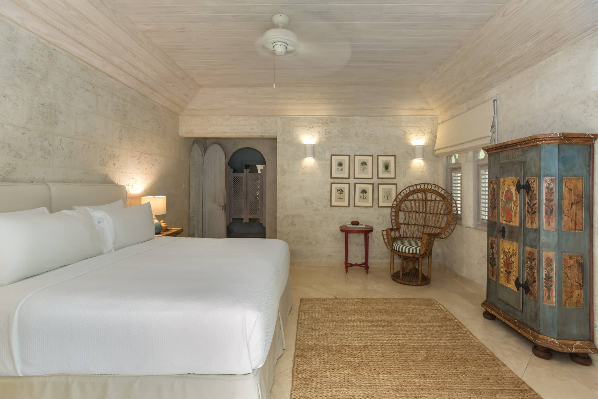 A cozy Bluff House and Cottage bedroom with a large white bed, wicker chair, rustic wardrobe with painted panels, framed art, woven rug, and neutral stone walls under a ceiling fan. Warm ambient lighting creates a calm atmosphere.