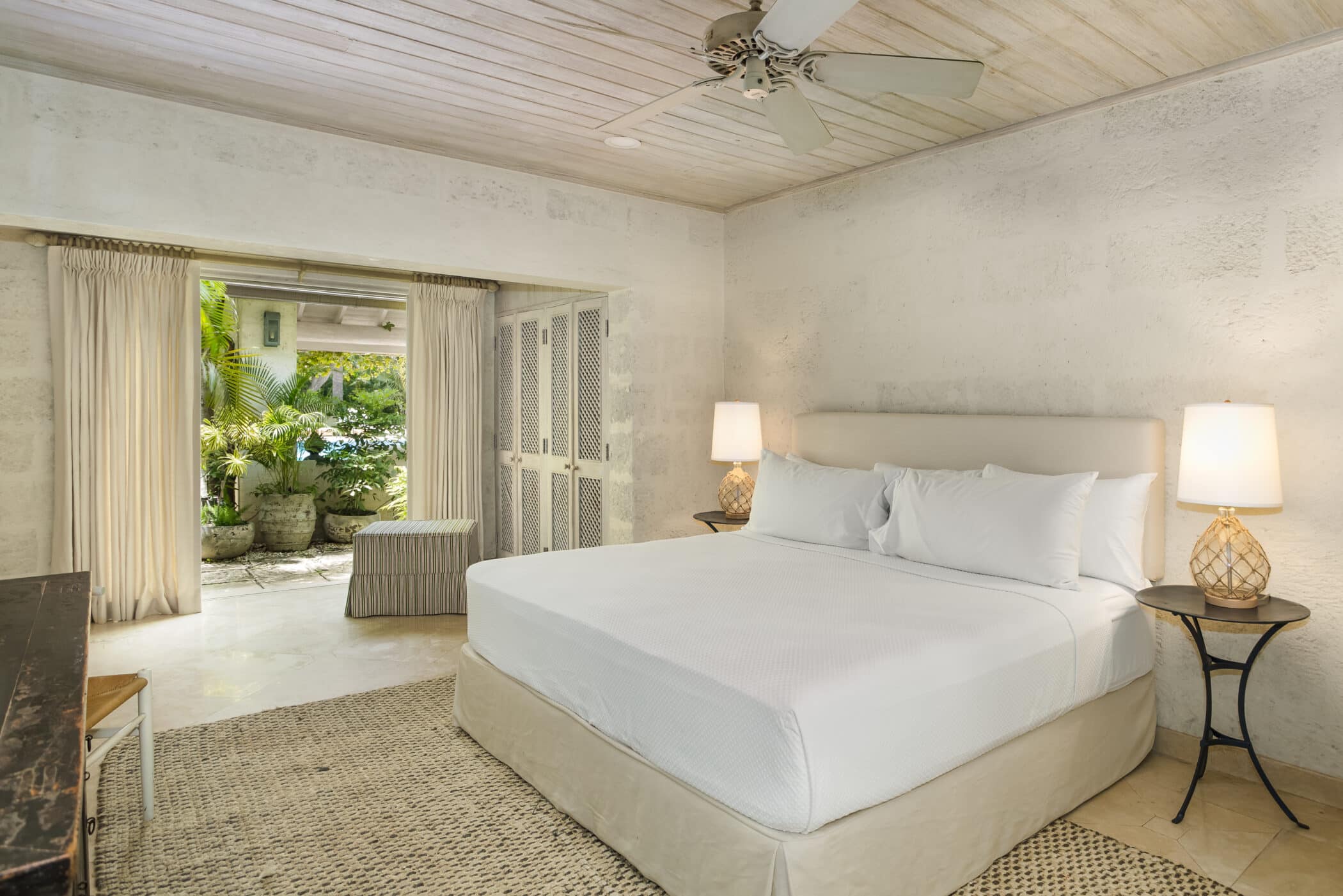 Bright, airy bedroom at Bluff House and Cottage with a large bed, two bedside lamps, woven rug, ceiling fan, and open doors leading to a lush garden patio with tropical plants.