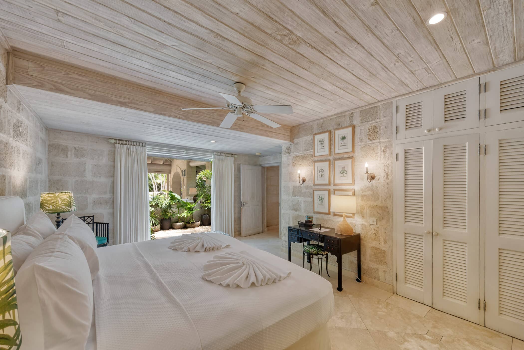 Bright, airy bedroom at Bluff House and Cottage with a white bed, ceiling fan, wooden ceiling, rustic stone walls, and framed art. Sliding doors open to a lush garden, while curtains and louvered closet doors enhance the tropical vibe.