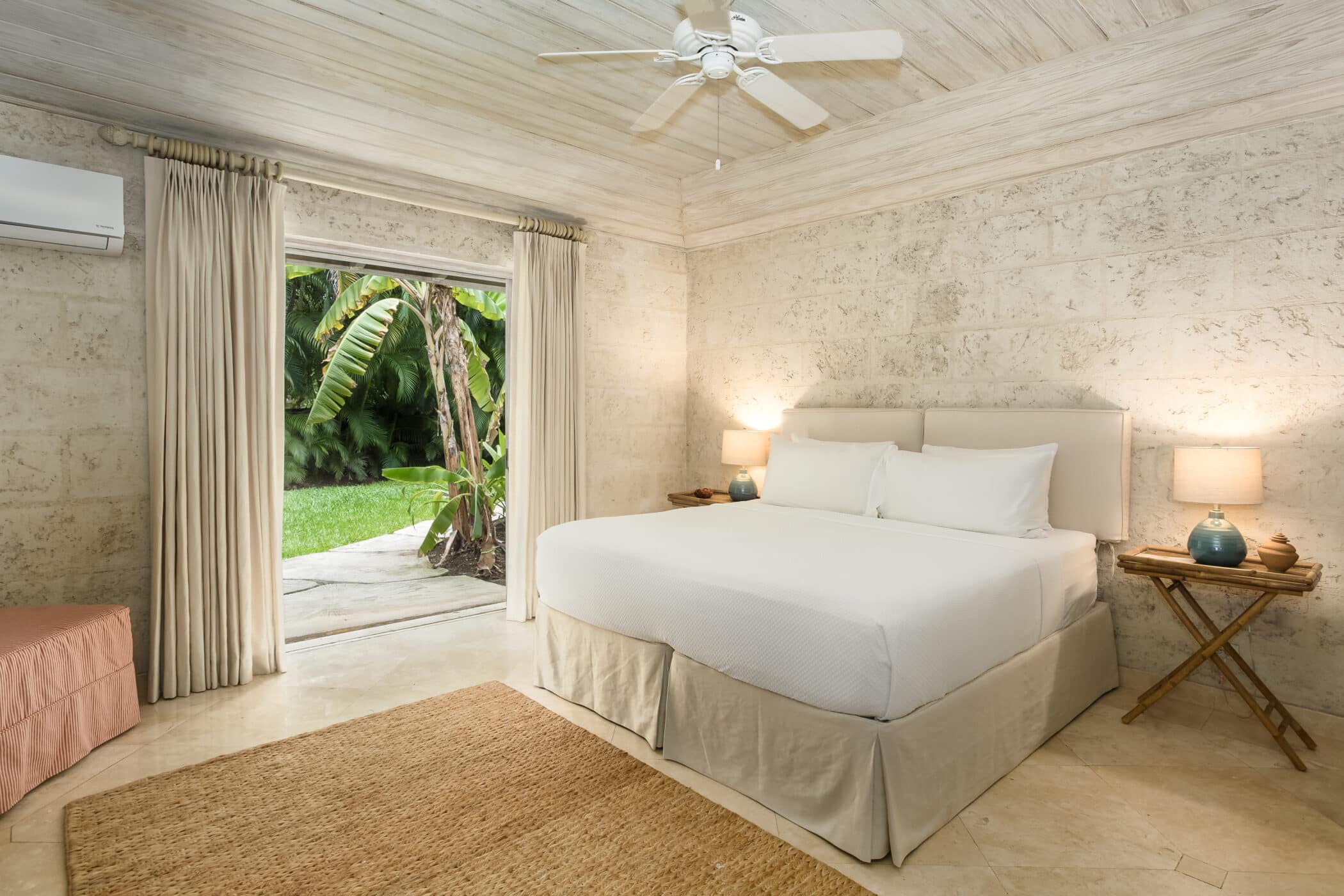 A bright bedroom at Bluff House and Cottage features stone walls, a large bed with white linens, two bedside tables with lamps, a ceiling fan, and an open door leading to a lush green garden.