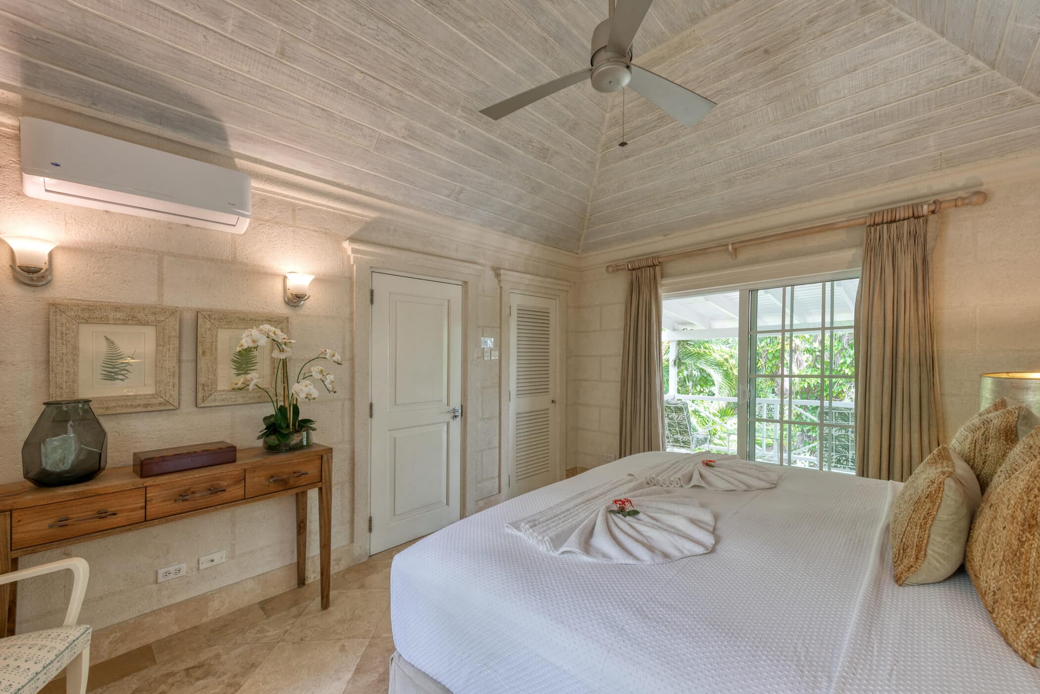 A bright, airy bedroom at Bluff House and Cottage features a king-sized bed, neutral decor, wooden ceiling with fan, and large windows with beige curtains letting in natural light. A wooden console table with decor sits against one wall.