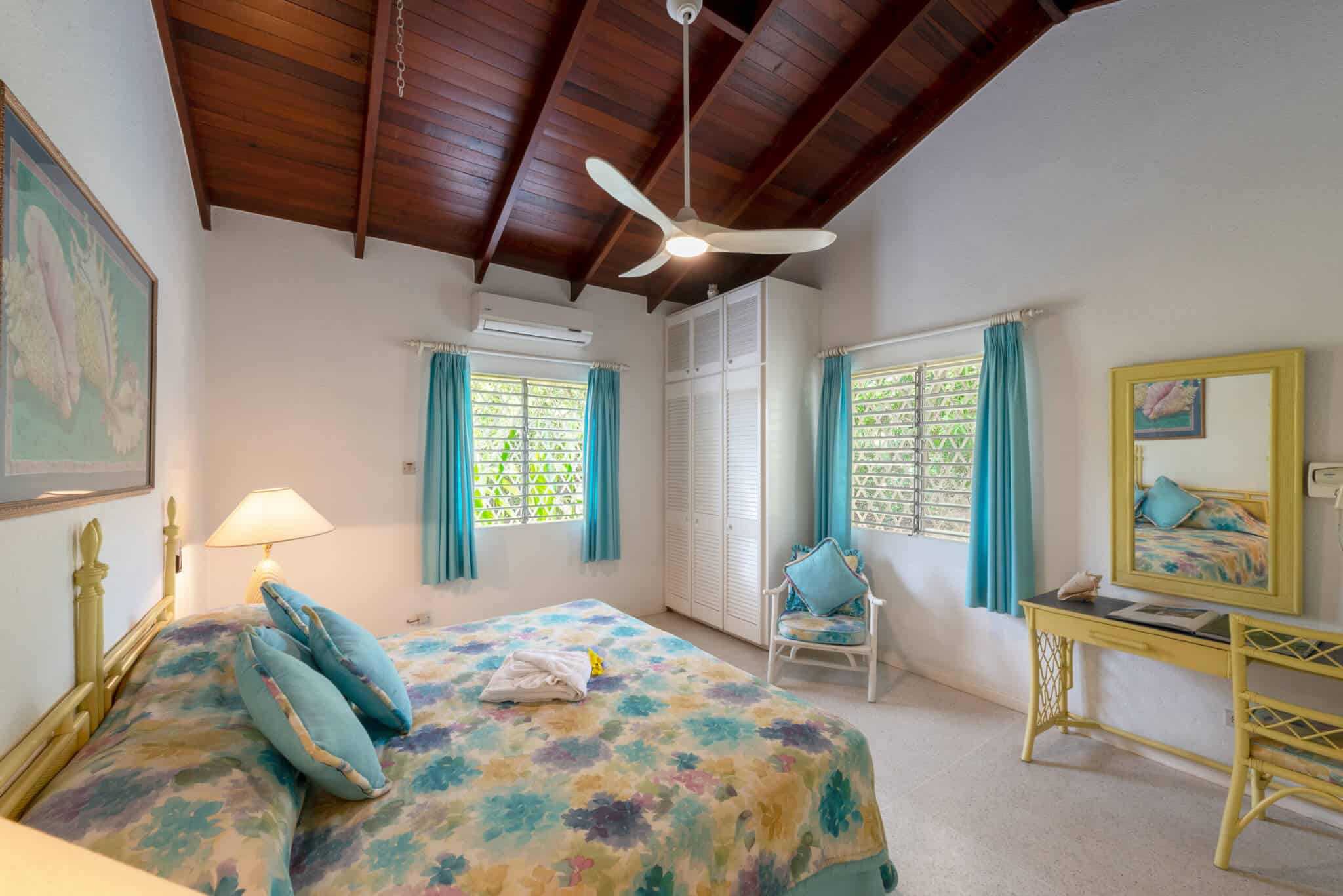 Bright Casabella bedroom with a floral bedspread, yellow furniture, large mirror, chair, two windows with blue curtains, a ceiling fan, air conditioner, lamp, and a wooden vaulted ceiling.