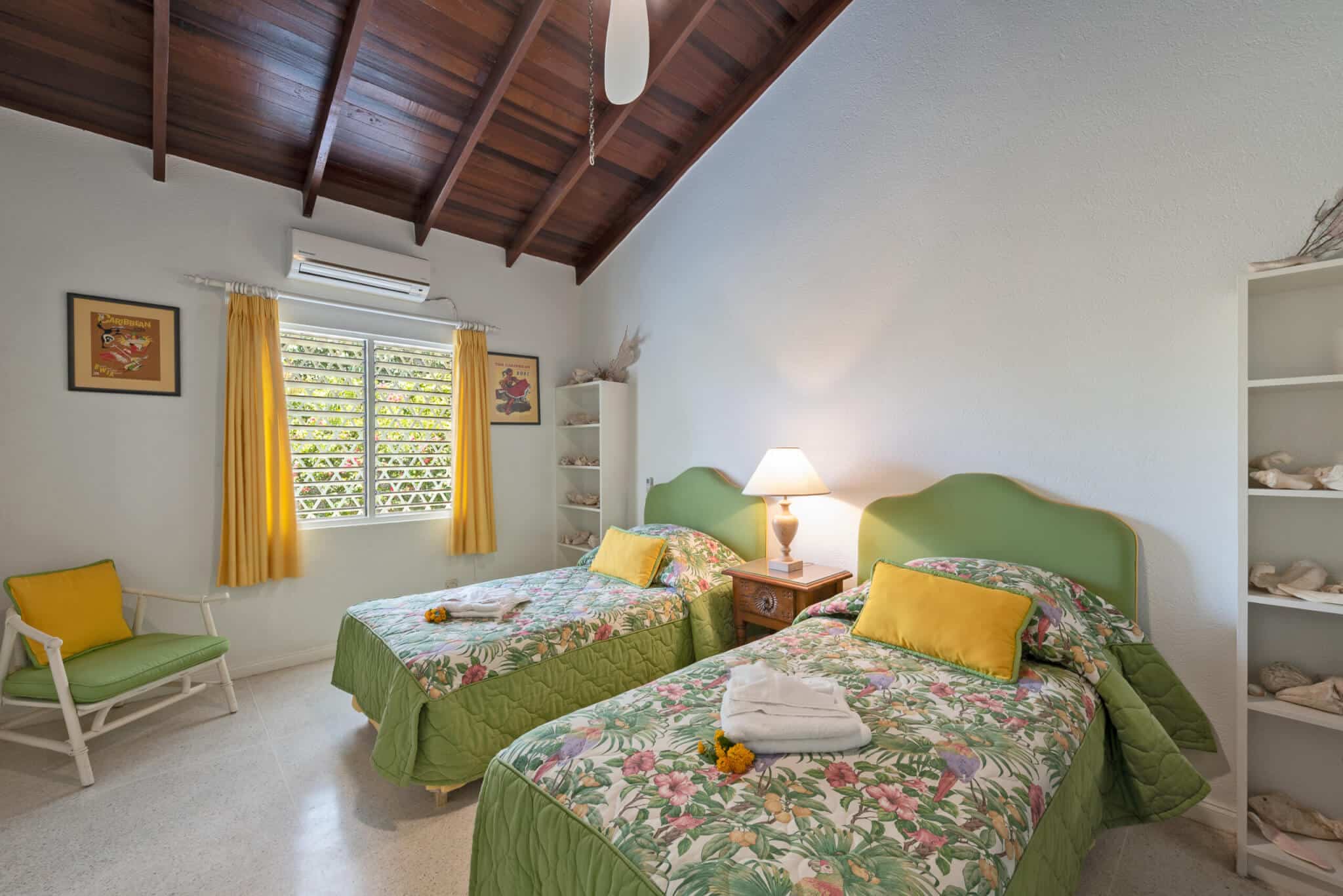 A bright Casabella bedroom with two twin beds featuring green floral bedding, yellow pillows, a lamp on a nightstand, white shelves with seashells, a green chair, and a window with yellow curtains under a wooden ceiling.