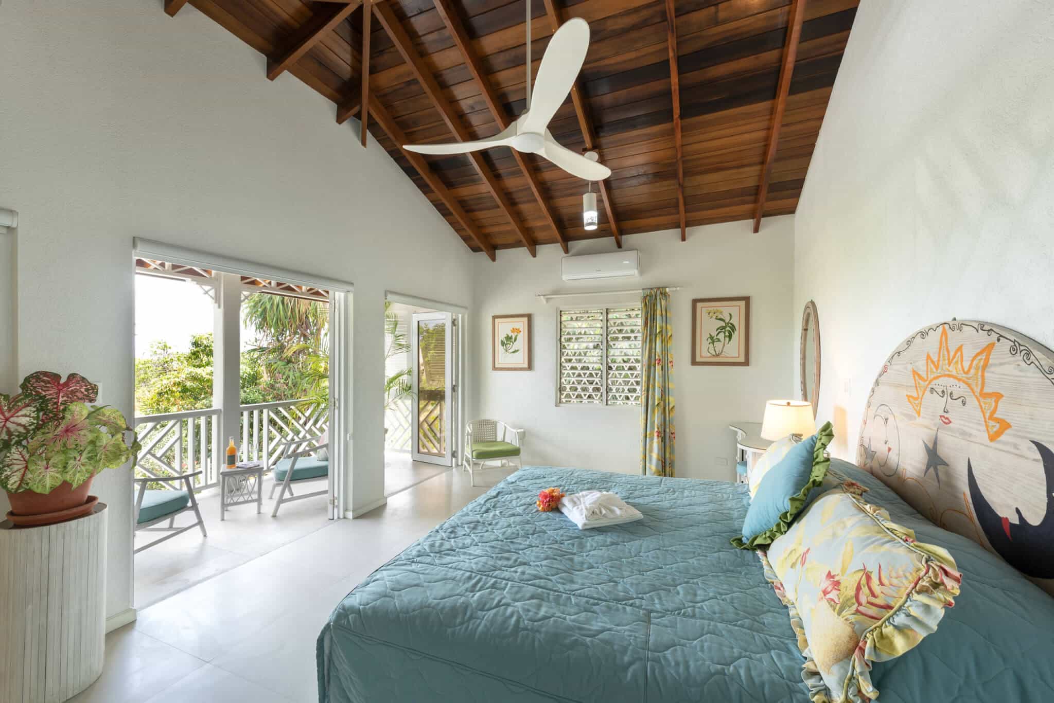 Bright Casabella bedroom with a vaulted wooden ceiling, large bed, tropical decor, ceiling fan, and open doors leading to a balcony with lounge chairs and lush greenery outside.