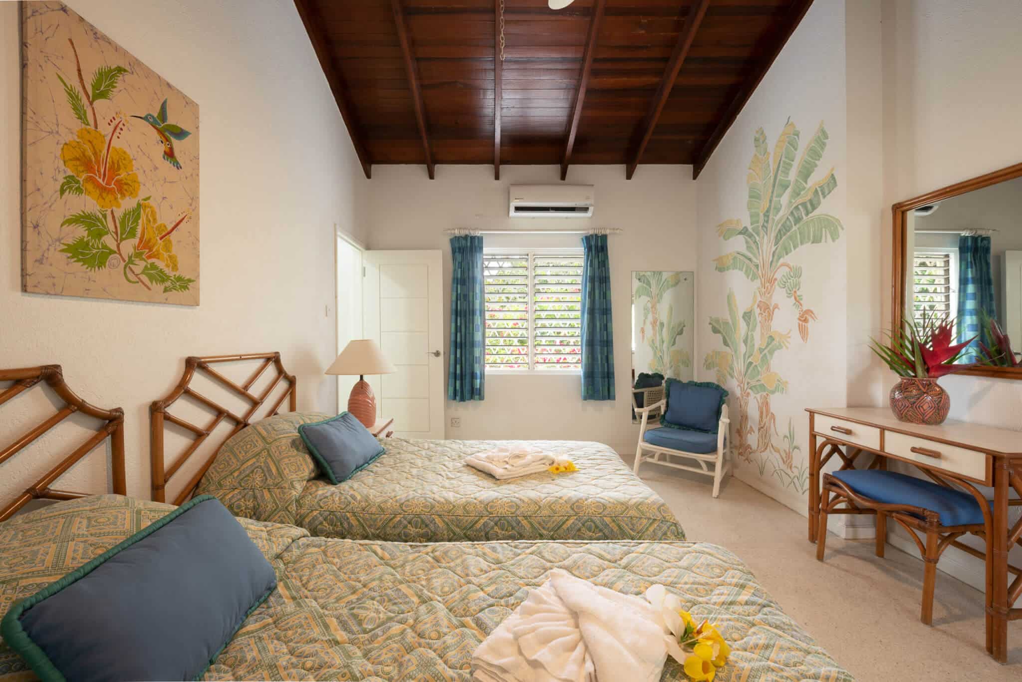 A bright Casabella bedroom with two twin beds, tropical-themed bedding, wall art, a chair, a desk with a mirror, and a window with blue curtains. Towels and yellow flowers are placed on the beds.