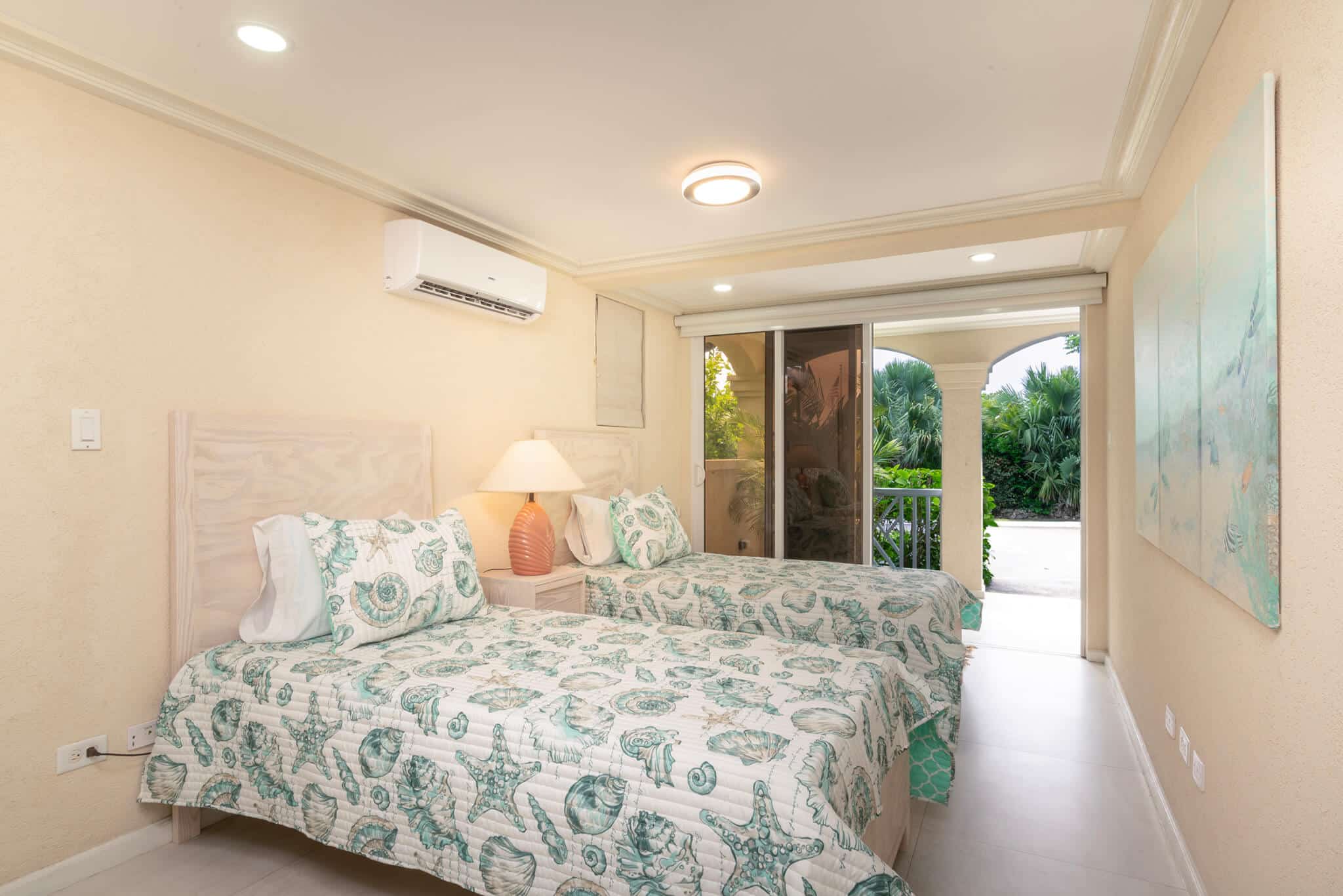 A bright Casabella bedroom with two twin beds featuring seashell-patterned bedding, a lamp on a nightstand, pastel wall art, an air conditioner, and glass doors opening to a lush, green patio area.