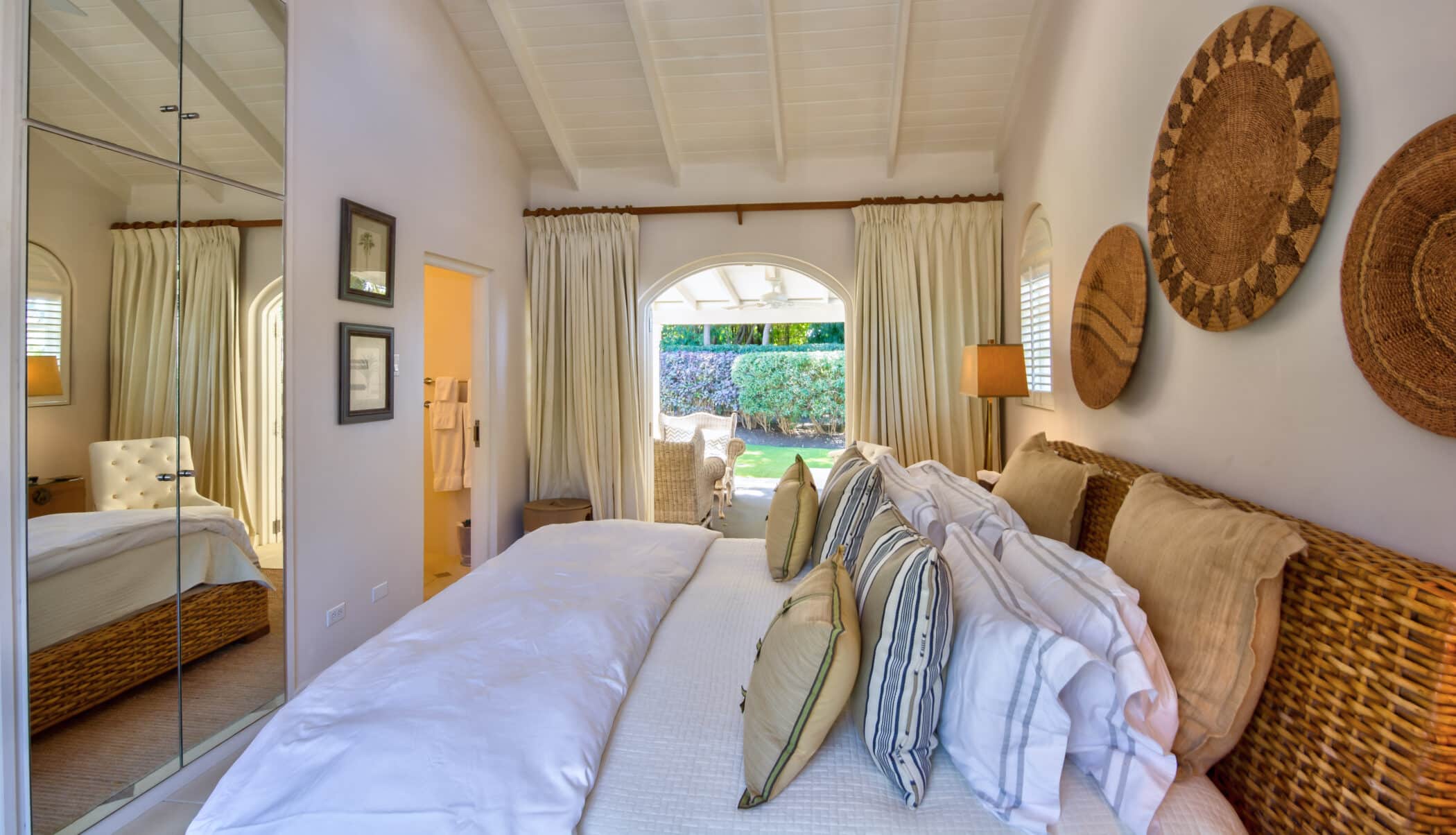 Bright bedroom with woven decor on the wall, a wicker headboard, white bedding, many pillows, mirrored closet doors, and open glass doors leading to an outdoor garden—experience the relaxed elegance of Casablanca at Sandy Lane.