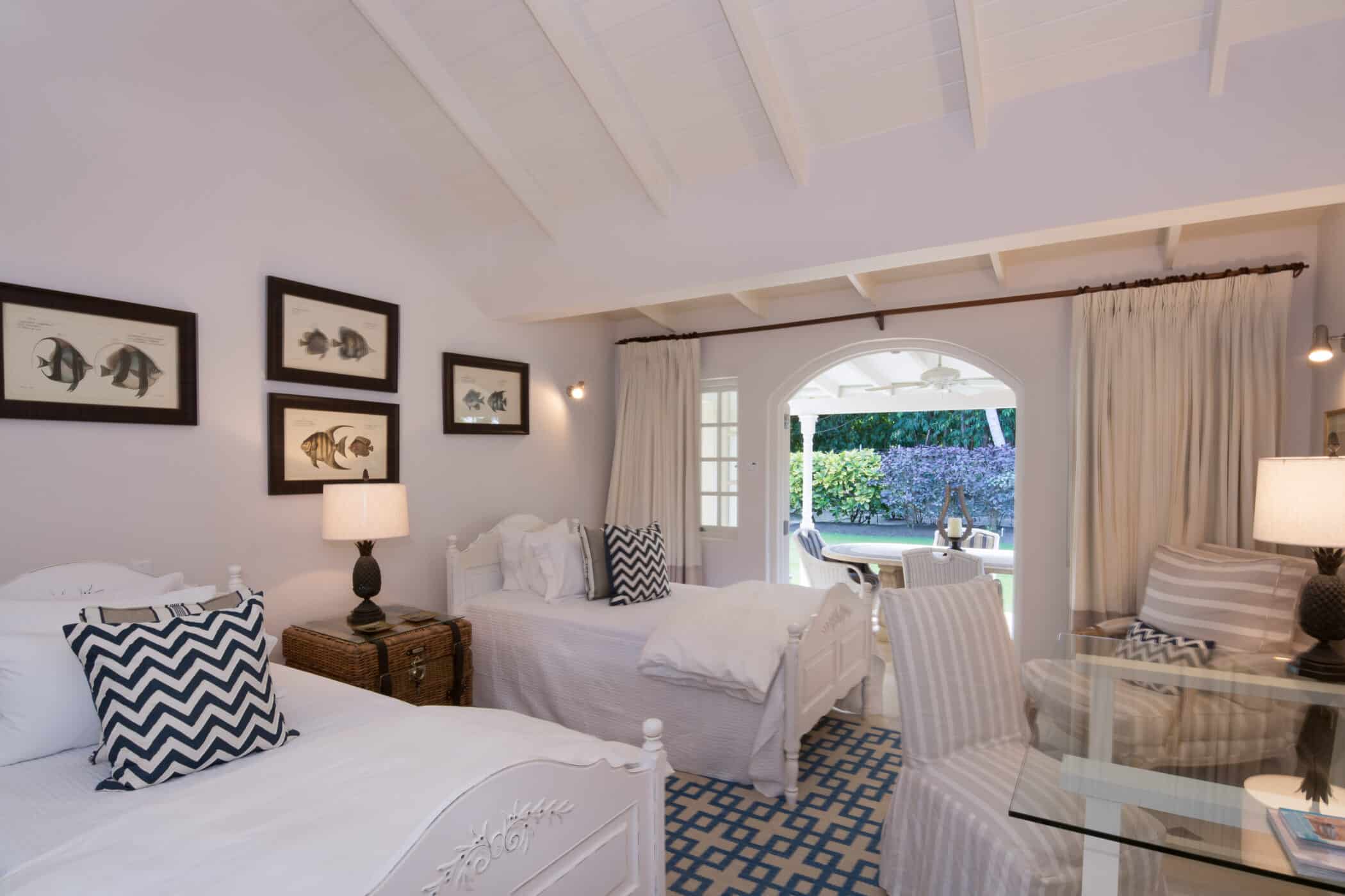 Bright bedroom at Casablanca at Sandy Lane with two white twin beds, chevron pillows, framed fish art, wicker nightstand, glass table, and sliding doors opening to a sunny garden patio with outdoor seating.