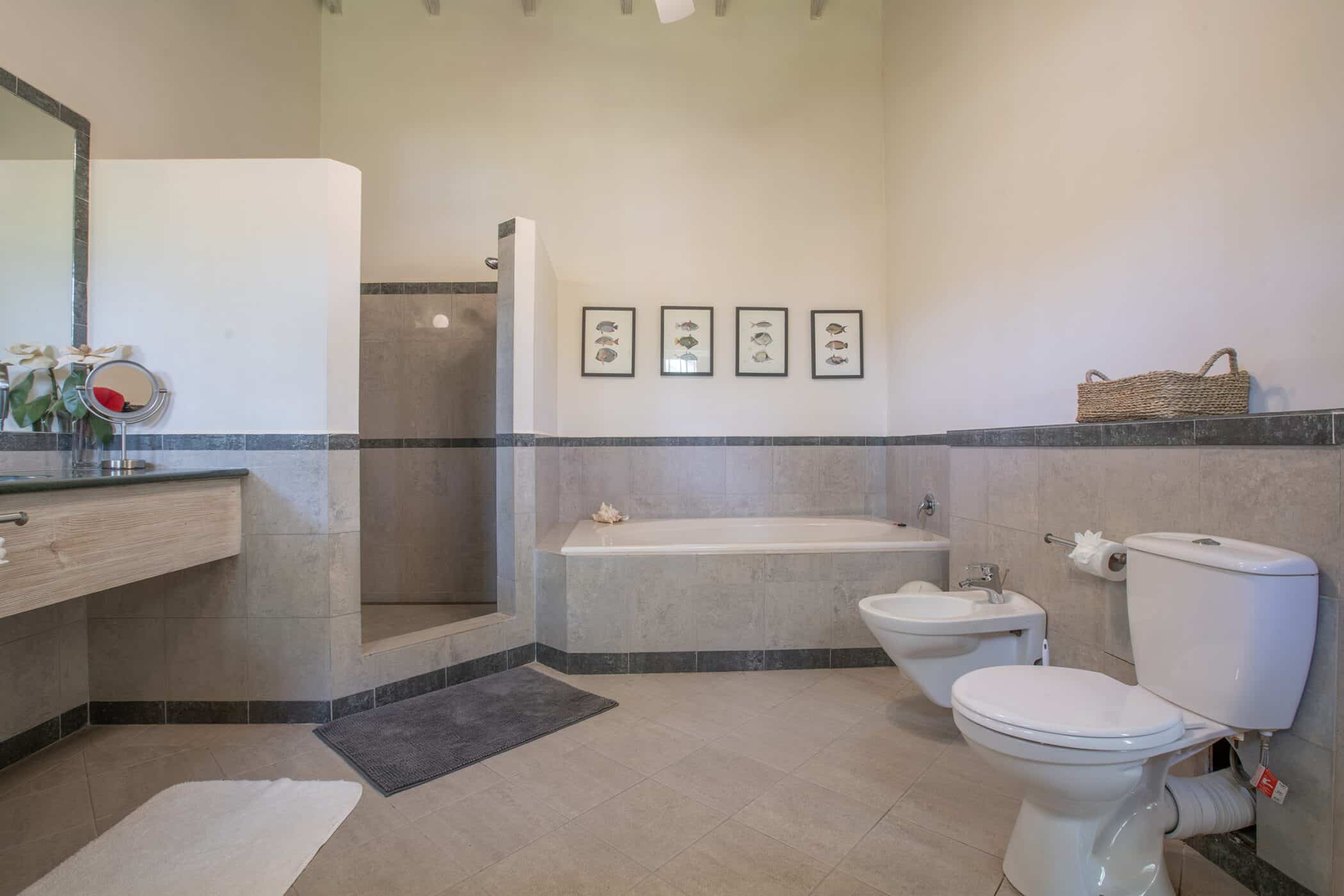 Spacious modern bathroom with tiled floors, a bathtub, walk-in shower, toilet, bidet, sink with a mirror, and decorative wall art above the tub. A basket and towels are placed on a shelf above the toilet.