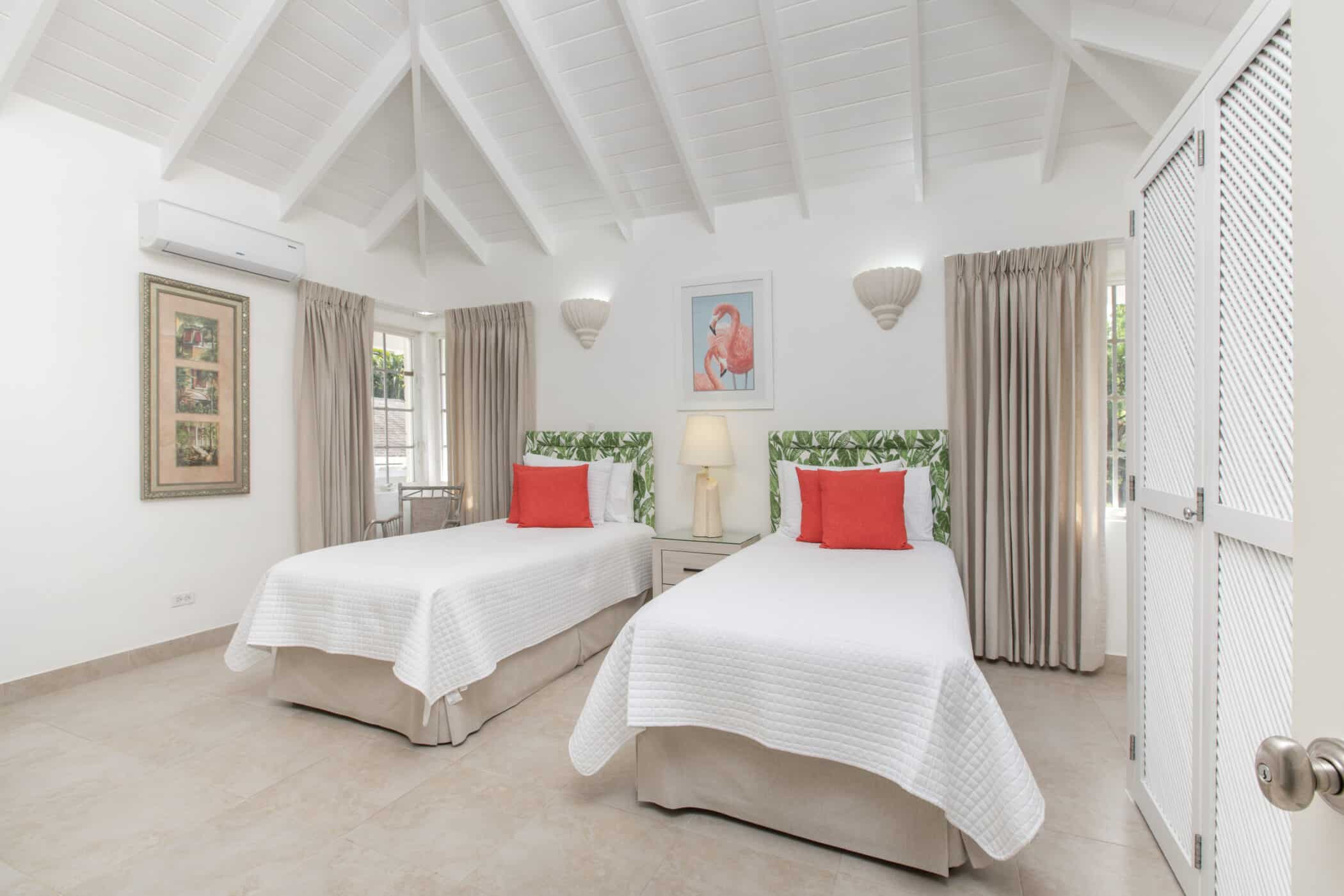 Bright bedroom with two single beds, white bedding, green and white Rose of Sharon headboards, red accent pillows, a lamp, framed artwork, beige curtains, and a vaulted white ceiling with exposed beams.