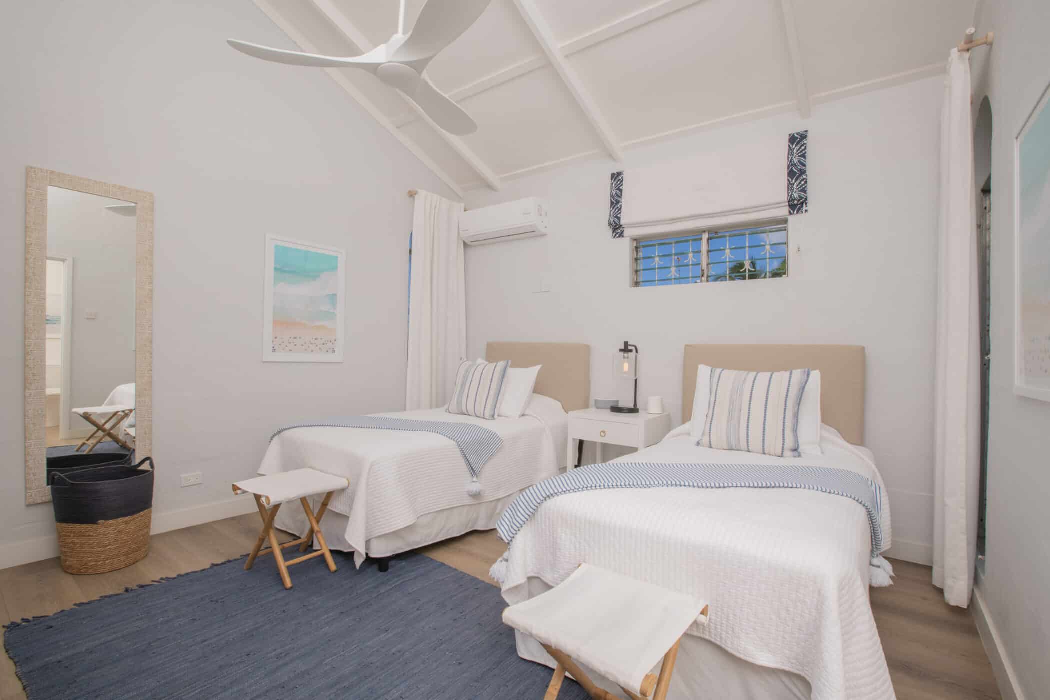 Bright bedroom with two single beds, blue rug, white walls, a ceiling fan, a large mirror, wicker basket, and a window with a valance. Bedside table and stools are placed beside the beds for added convenience. This inviting space is part of Secret Cove Two, offering comfort and coastal charm for your stay.
